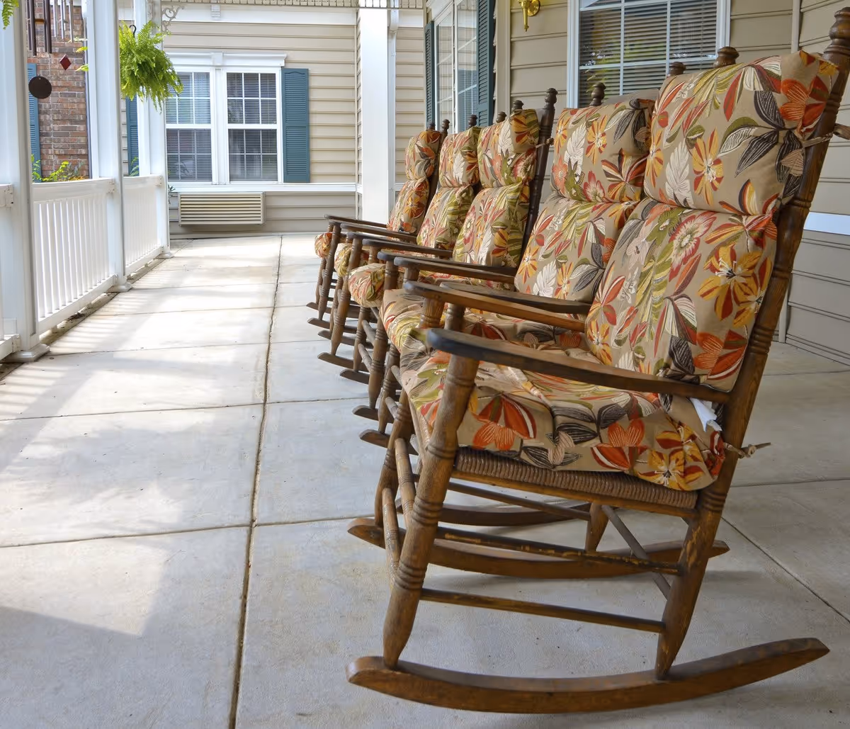 A row of wooden rocking chairs with floral patterned cushions lined up on a covered porch with concrete flooring. The porch has white railings and is attached to a building with beige siding and windows with white trim and blue shutters.