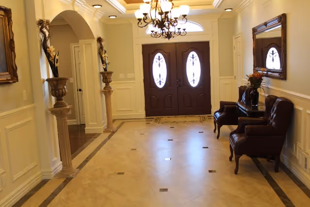 A spacious and elegant entryway with double dark wooden doors featuring oval glass panels. The floor is tiled with a beige and brown pattern. On the right side, there are two dark leather armchairs with a small table in between, decorated with a vase of flowers. A large mirror hangs above the chairs. On the left side, there are two tall decorative pedestals with floral arrangements. The ceiling has a chandelier and recessed lighting, and the walls are painted a soft yellow with white wainscoting.