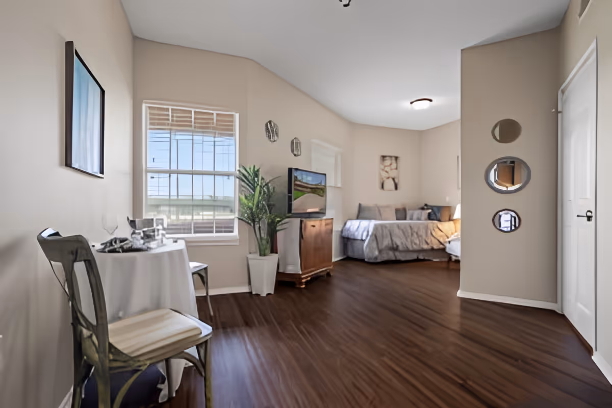 A bright and spacious bedroom with a bed, a small dining table with two chairs, a TV on a wooden stand, a potted plant, and three decorative round mirrors on the wall near a closed door. The room has wooden flooring and beige walls with a window letting in natural light.