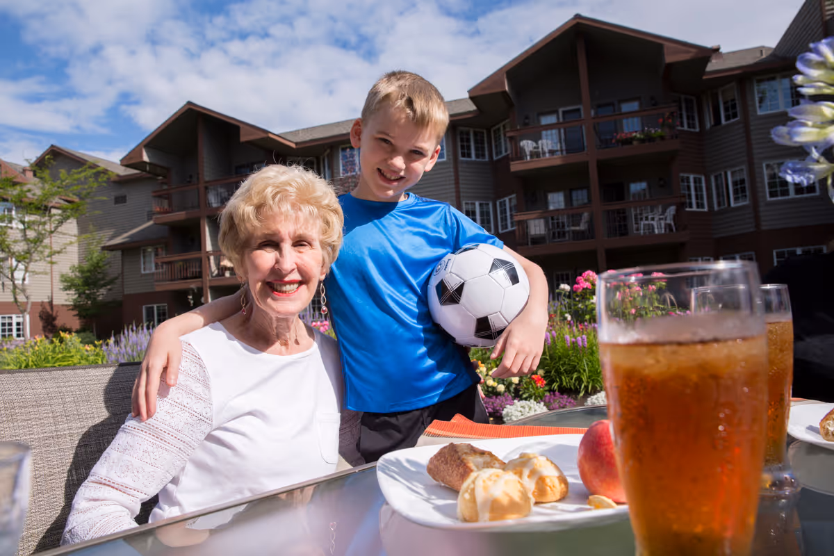 An elderly woman and a young boy smiling outdoors at a senior living facility. The boy is holding a soccer ball and has his arm around the woman. They are sitting at a glass table with plates of food and glasses of iced tea. Behind them is a multi-story building with balconies and a garden with flowers.