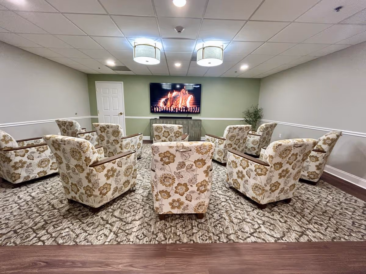 A cozy common area with eight floral-patterned armchairs arranged in a semi-circle facing a wall-mounted flat-screen TV displaying a fireplace scene. The room has a green accent wall, white door, two ceiling lights, and a patterned carpet.