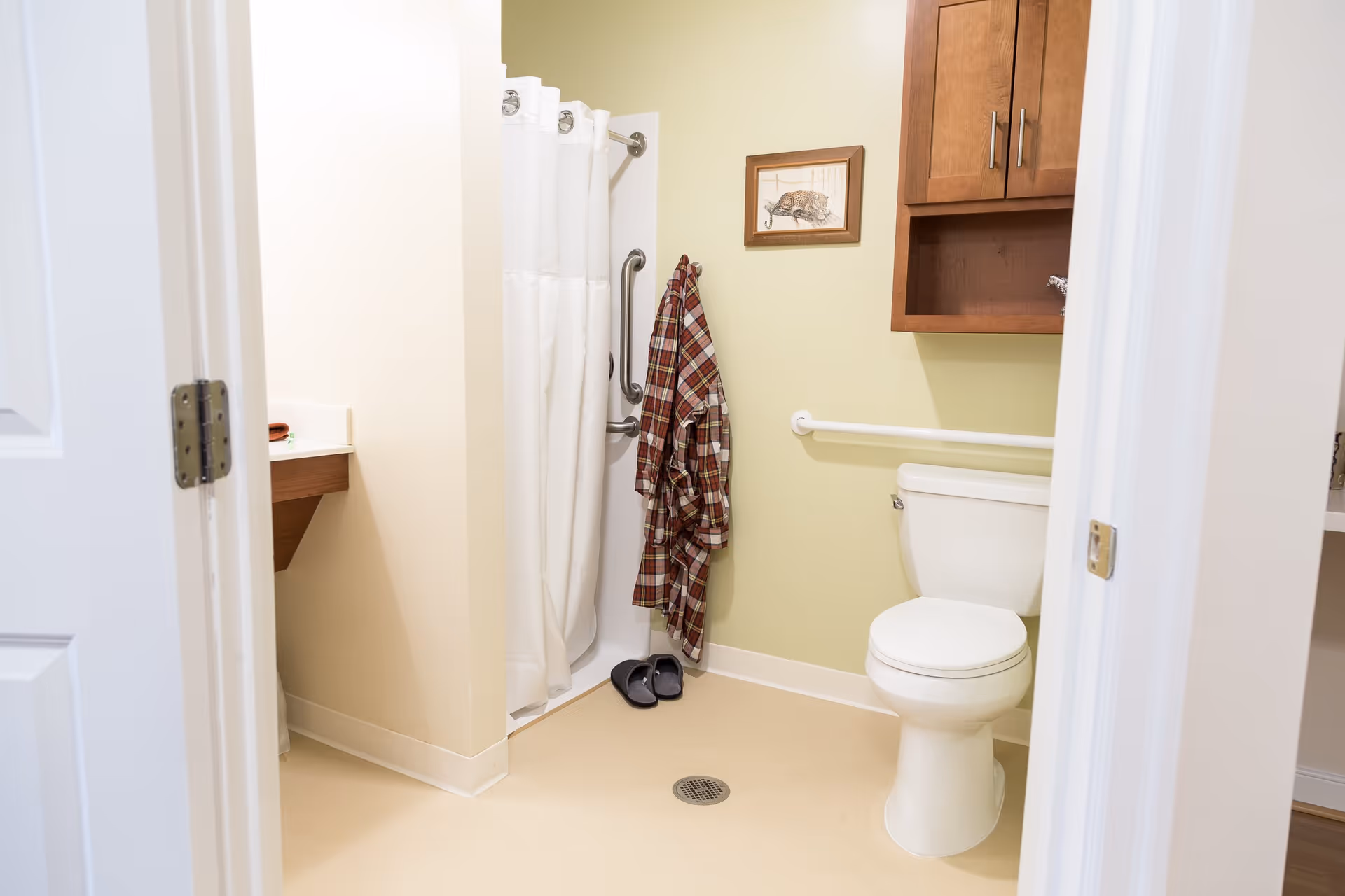 A clean and accessible bathroom with a white toilet, a shower with a white curtain, a wooden cabinet mounted on the wall, a plaid robe hanging on a hook, and a pair of slippers on the floor. The walls are painted light green and beige, and there is a framed picture of a cat above the robe.