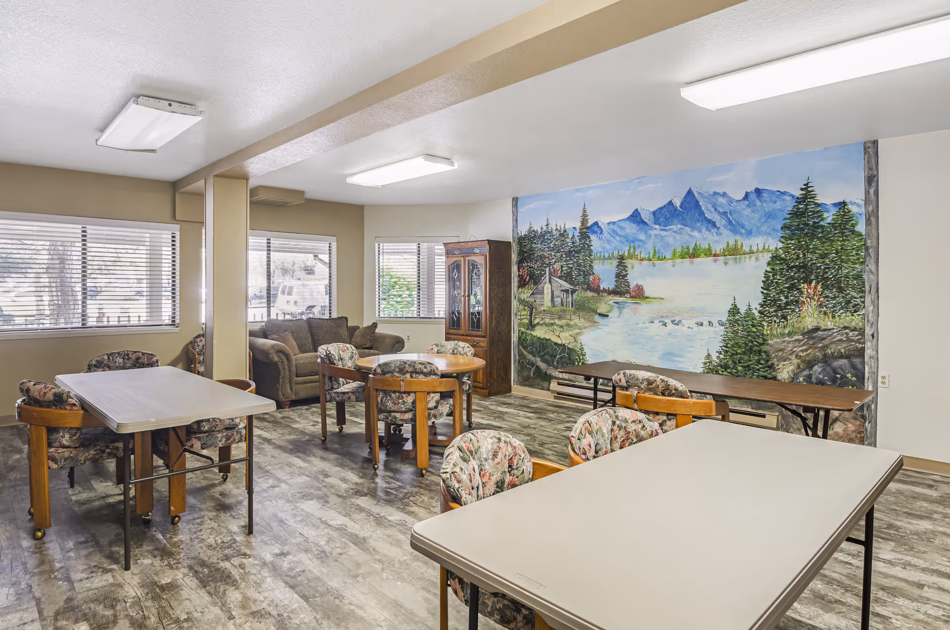 A communal room with multiple tables and floral cushioned chairs, a brown couch near windows with blinds, and a large mural on the wall depicting a scenic mountain lake with trees and a cabin.