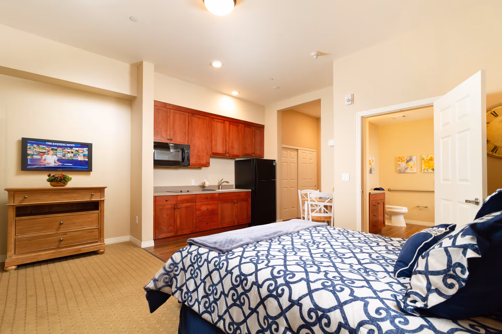 Interior view of a senior living facility room at Westmont of Chico featuring a bed with blue and white patterned bedding in the foreground, a wooden dresser with a flat-screen TV mounted above it on the left wall, a kitchenette with wooden cabinets, a microwave, and a black refrigerator in the background, and an open door leading to a bathroom with a toilet and floral wall art.