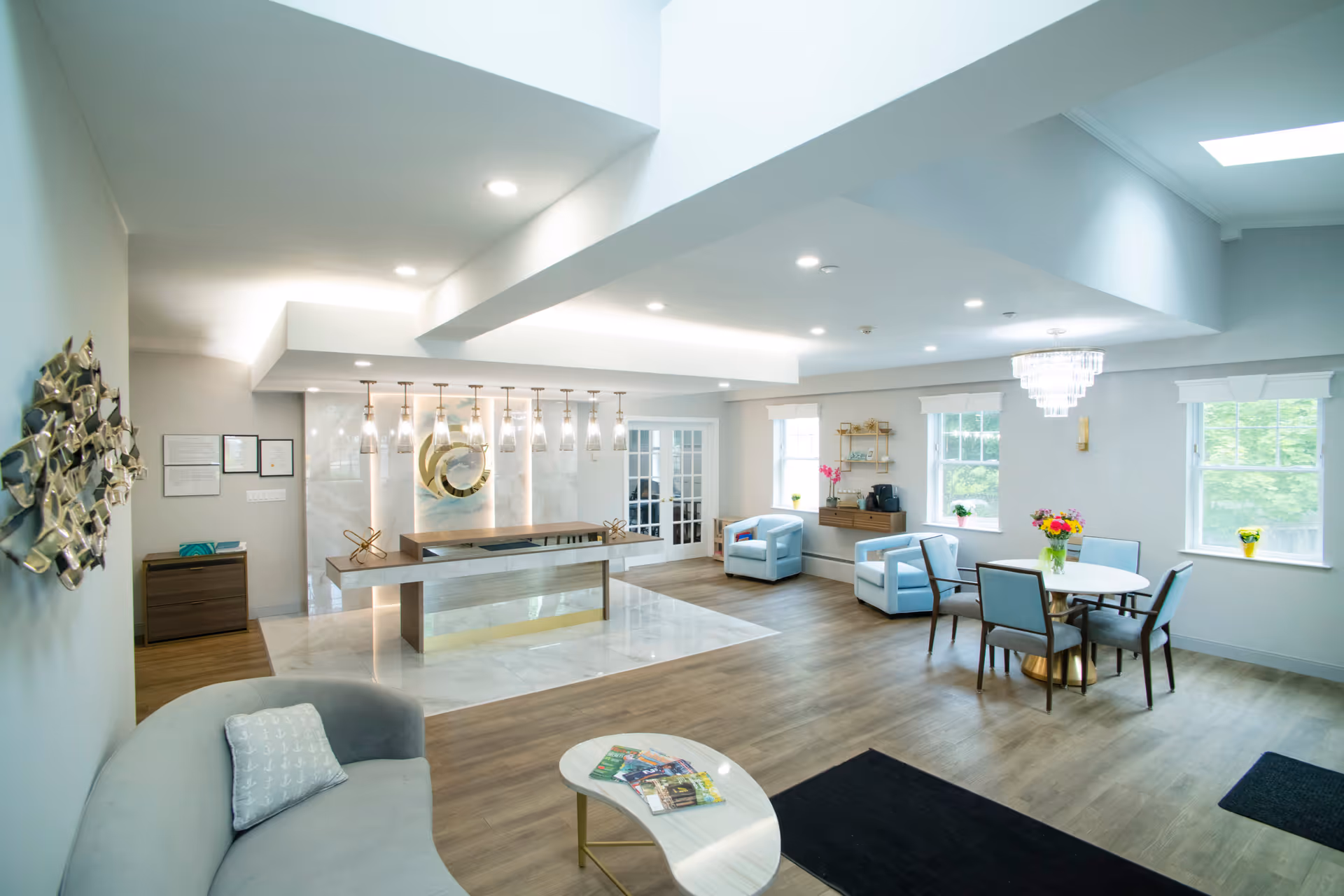 Bright and modern common area with a curved gray sofa, a small white marble coffee table with magazines, a reception desk with hanging pendant lights, two light blue armchairs, a round dining table with four chairs, large windows letting in natural light, and decorative wall art and flowers.