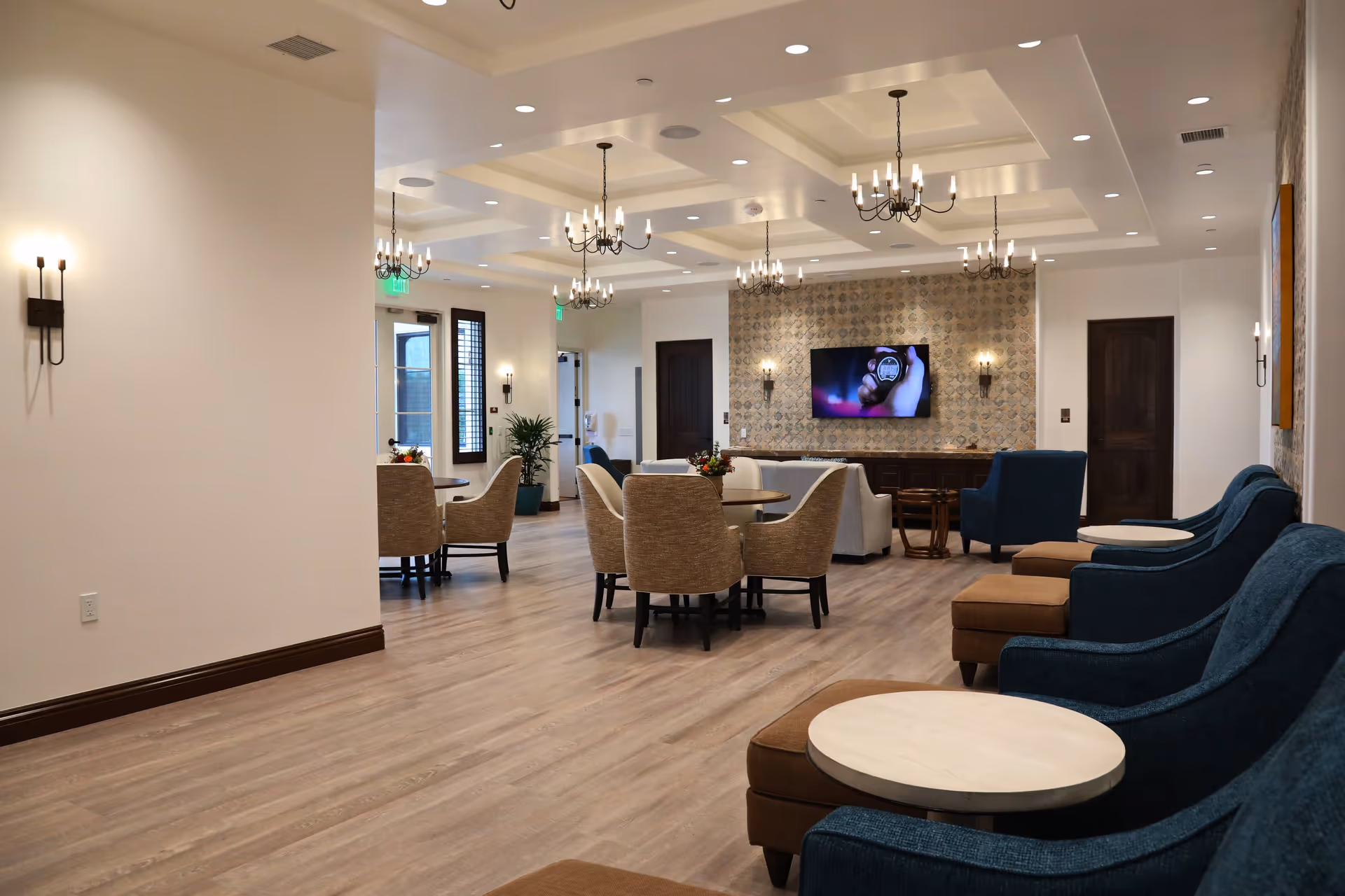 A spacious and well-lit living room area with multiple seating options including blue armchairs with round side tables and beige upholstered chairs around circular tables. The room features a patterned accent wall with a mounted flat-screen TV, elegant chandeliers hanging from a coffered ceiling, and wall sconces providing additional lighting. The floor is covered with light wood laminate, and there are large windows and doors letting in natural light.