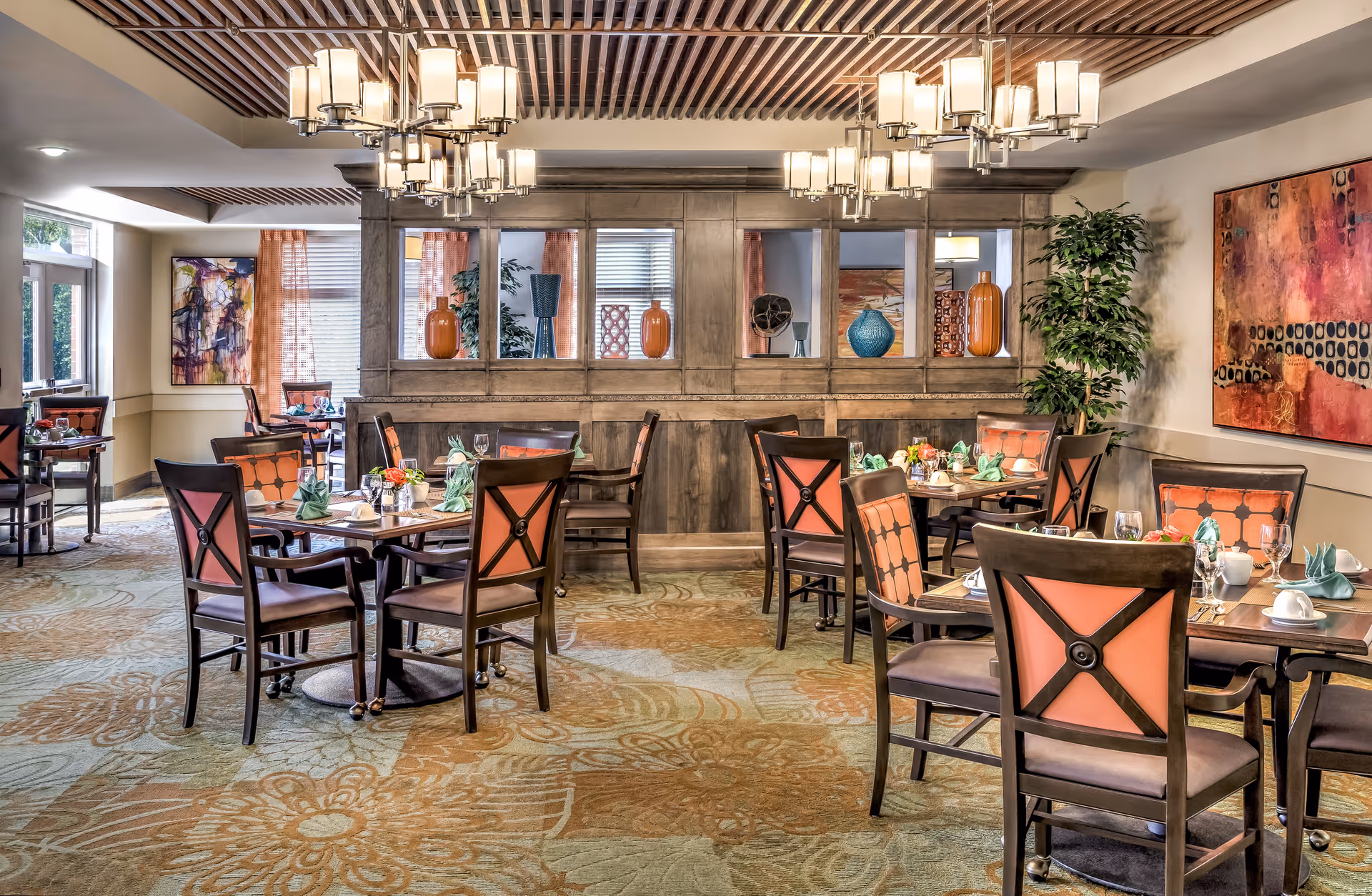 A well-lit dining room with several wooden tables and chairs arranged neatly. Each table is set with green folded napkins, glassware, and small floral centerpieces. The room features a patterned carpet, decorative vases on a wooden partition, large abstract paintings on the walls, and modern chandeliers hanging from a wooden slatted ceiling.