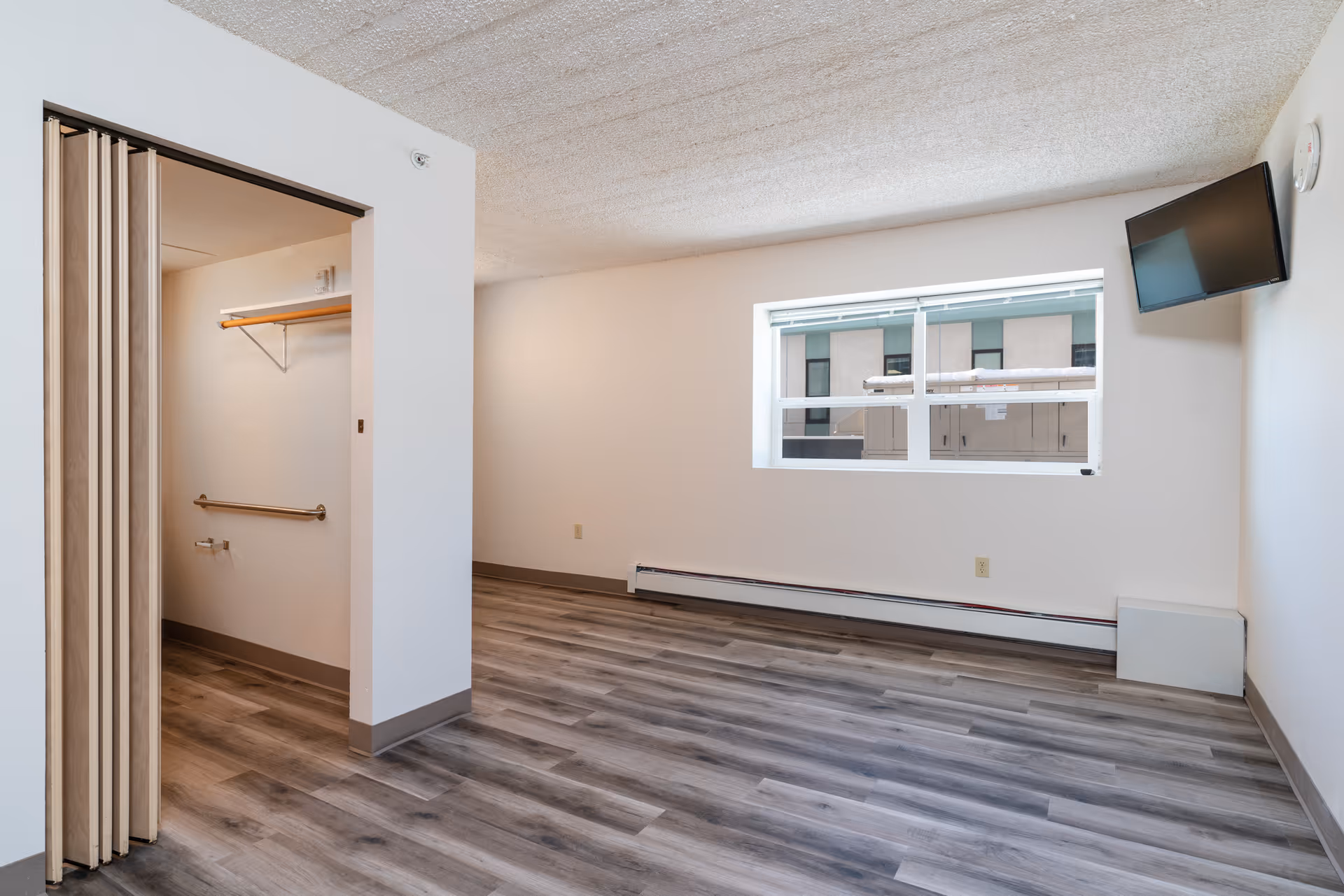 Empty room with wood-style flooring, a window showing an exterior building, a wall-mounted TV, and an open closet with a folding door and a shelf inside.