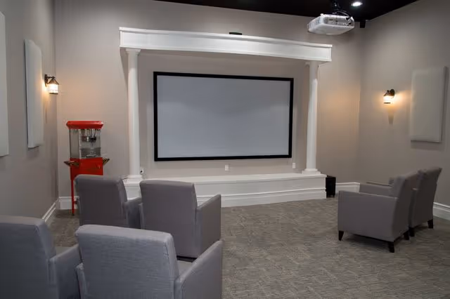 A small theater room with a large blank projection screen framed by white columns. There are six gray upholstered armchairs arranged in two rows facing the screen. A red popcorn machine is positioned in the back left corner of the room. The walls are light gray with wall-mounted lights and soundproof panels. A ceiling-mounted projector is visible above the seating area.
