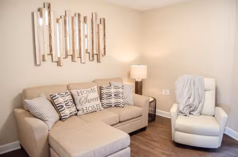 A cozy living room area with a beige sectional sofa adorned with several decorative pillows, including one that says 'Home Sweet Home.' Next to the sofa is a white recliner chair draped with a light gray blanket. A modern table lamp on a small side table provides warm lighting. The walls are light-colored, and a decorative mirror with vertical rectangular panels hangs above the sofa. The floor is wooden.