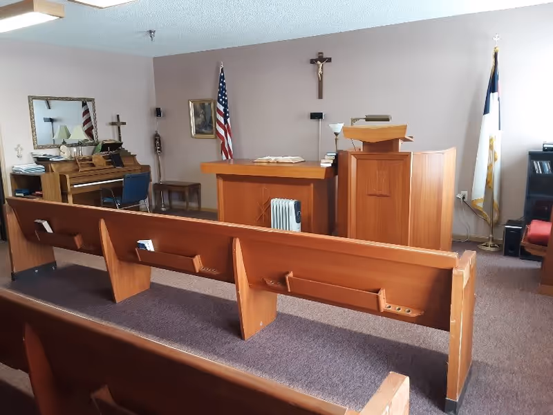 Interior view of a small chapel or prayer room with wooden pews, a wooden altar and lectern, an American flag and a Christian flag, a crucifix on the wall, a piano with a chair, and religious decorations.