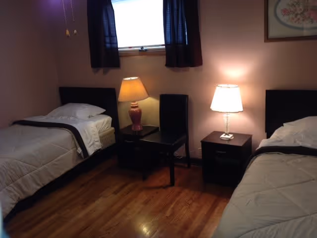A bedroom with two single beds, each with white bedding and a pillow. Between the beds are two small dark wooden nightstands, each with a lamp on top. A black chair is placed between the nightstands. A window with dark curtains is above the nightstands, and a framed picture hangs on the wall to the right.