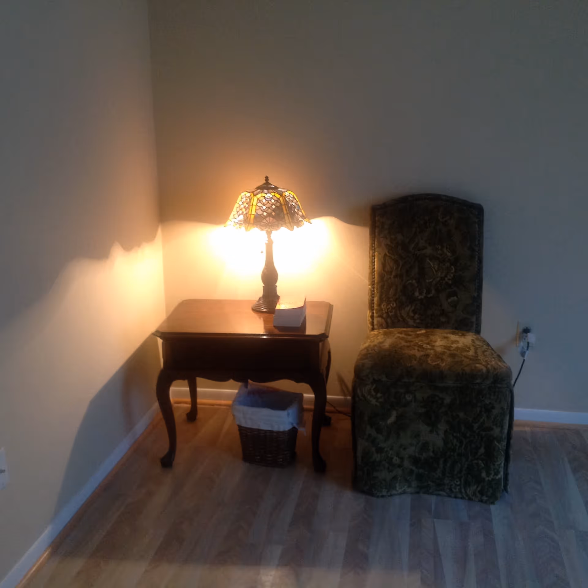 A small wooden side table with curved legs holding a lit stained glass table lamp and a box of tissues. Next to the table is a green upholstered chair with a floral pattern. The floor is light-colored wood, and the walls are plain and light-colored.