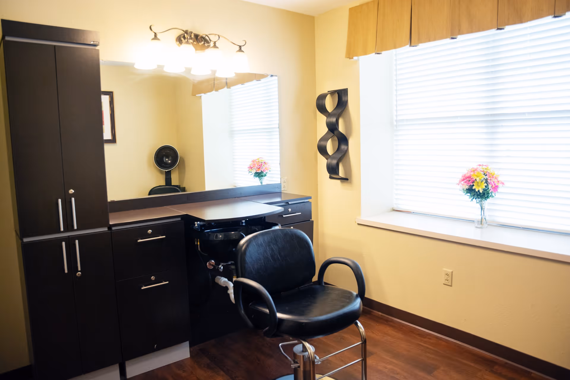 A well-lit hair salon station in a senior living facility with a black salon chair, a large mirror, dark wood cabinets, and a window with blinds and a vase of colorful flowers on the windowsill.
