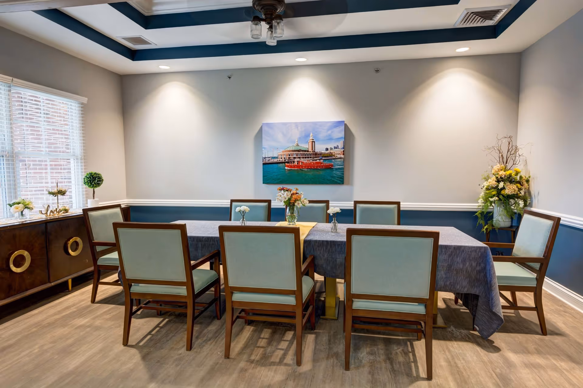 A dining room with a rectangular table covered with a gray tablecloth and surrounded by eight wooden chairs with light green cushions. The room has light gray walls with a blue accent wall and white trim. A large window with blinds is on the left side, and a sideboard with decorative items is beneath it. A colorful painting of a waterfront scene hangs on the wall above the table. There are floral arrangements on the table and in the corner of the room. The ceiling has a blue tray design with a ceiling fan and light fixture.