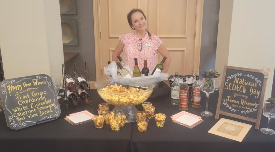 A woman stands behind a table set up for a happy hour event with various bottles of wine and whiskey, a large bowl of popcorn, and small cups filled with popcorn. Two chalkboard signs on either side of the table list happy hour wines and National Scotch Day featuring James Alexander. The setting appears to be indoors with a wooden door in the background.