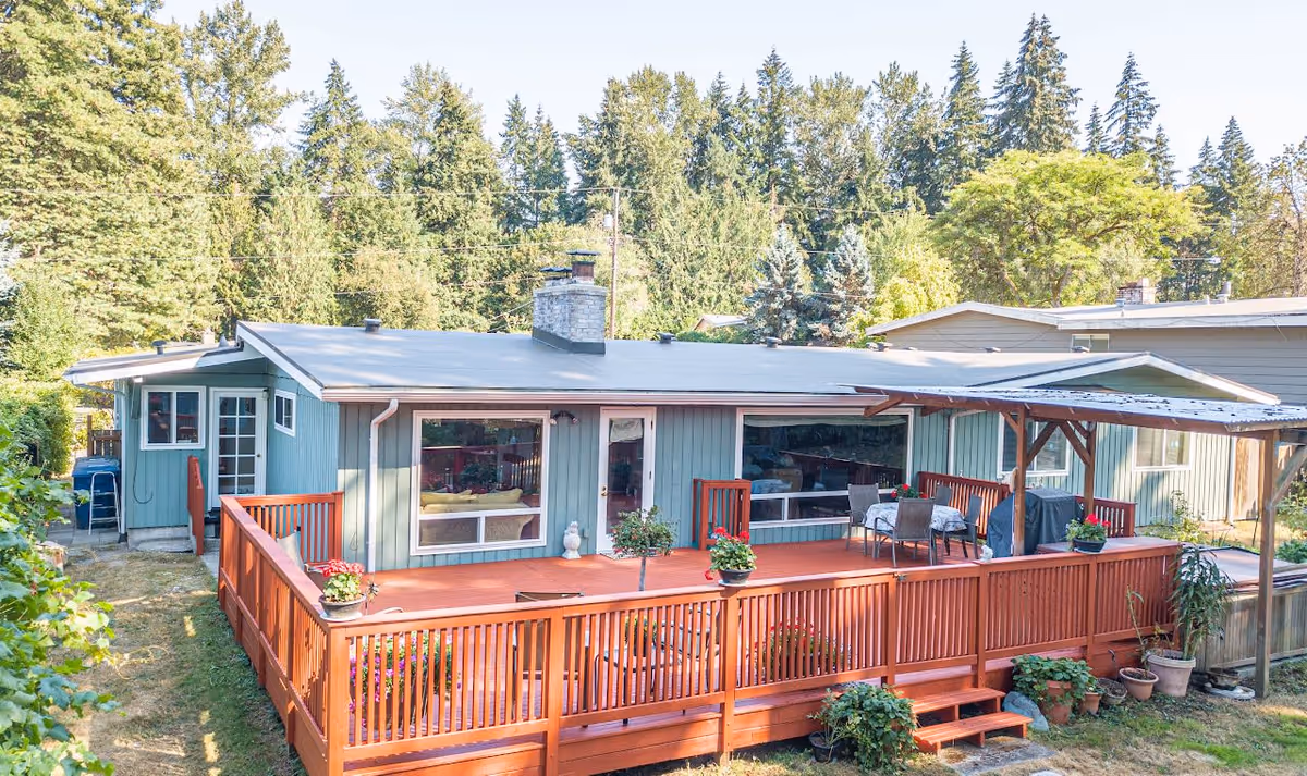 Exterior view of a single-story residential building with light blue siding and a flat roof. The building features large windows and a wooden deck painted in reddish-brown, with potted plants and outdoor furniture including a table and chairs under a pergola. The surrounding area has green trees and a grassy yard.