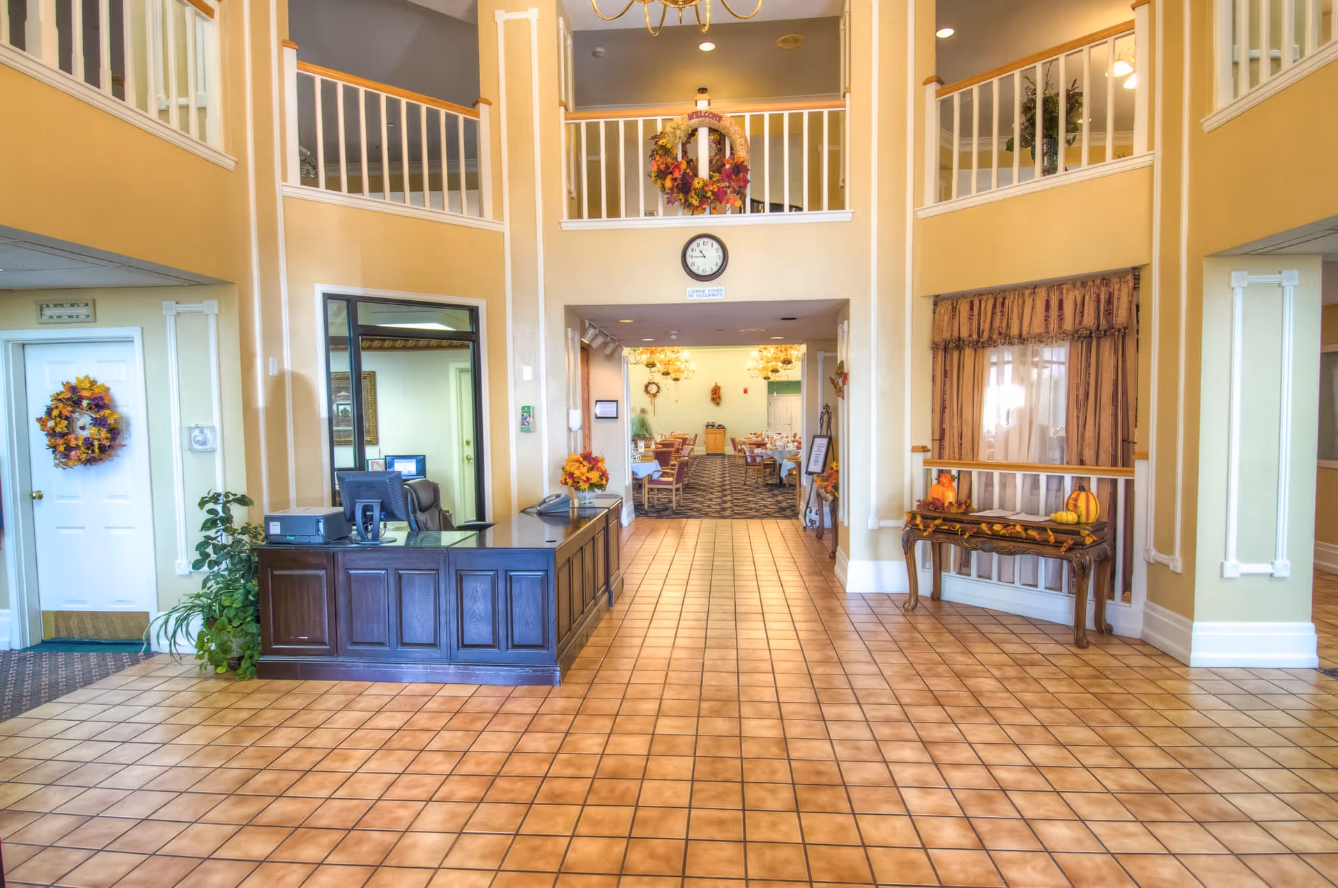 Spacious tiled lobby with a dark wood reception desk, decorative table and fall wreaths, and a view into a dining area beneath a two-story balcony.