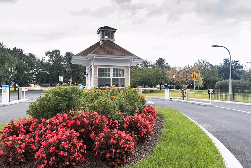 Gatehouse at the entrance to a senior living community with red flowering beds, a driveway, and surrounding trees.
