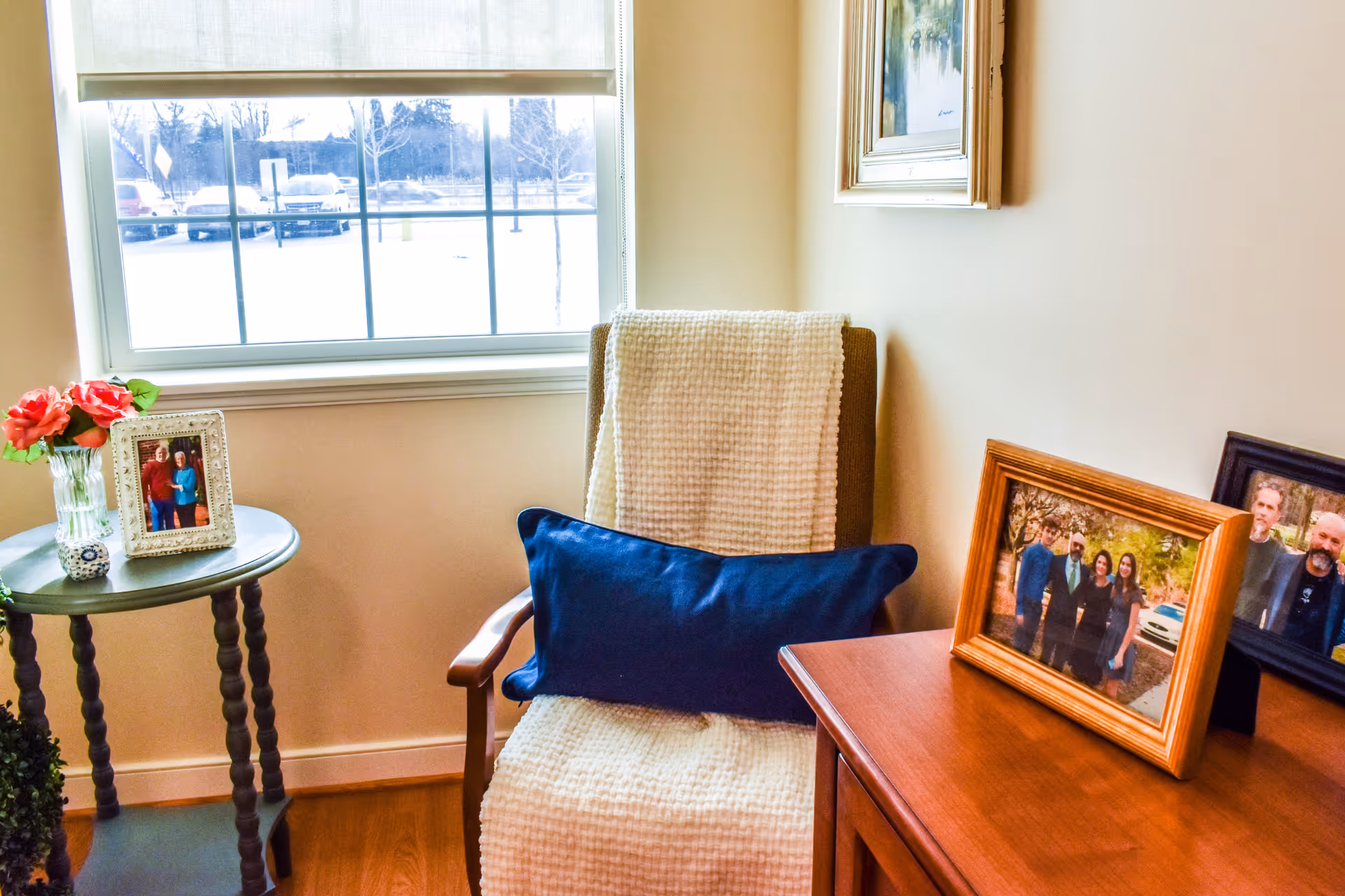 A cozy corner of a room with a wooden armchair draped with a cream-colored textured blanket and a dark blue pillow. Next to the chair is a small round table holding a vase with pink flowers and a framed photo. On the right side, there is a wooden dresser with two framed family photos. A window with a beige roller shade shows a snowy outdoor scene with parked cars and trees.