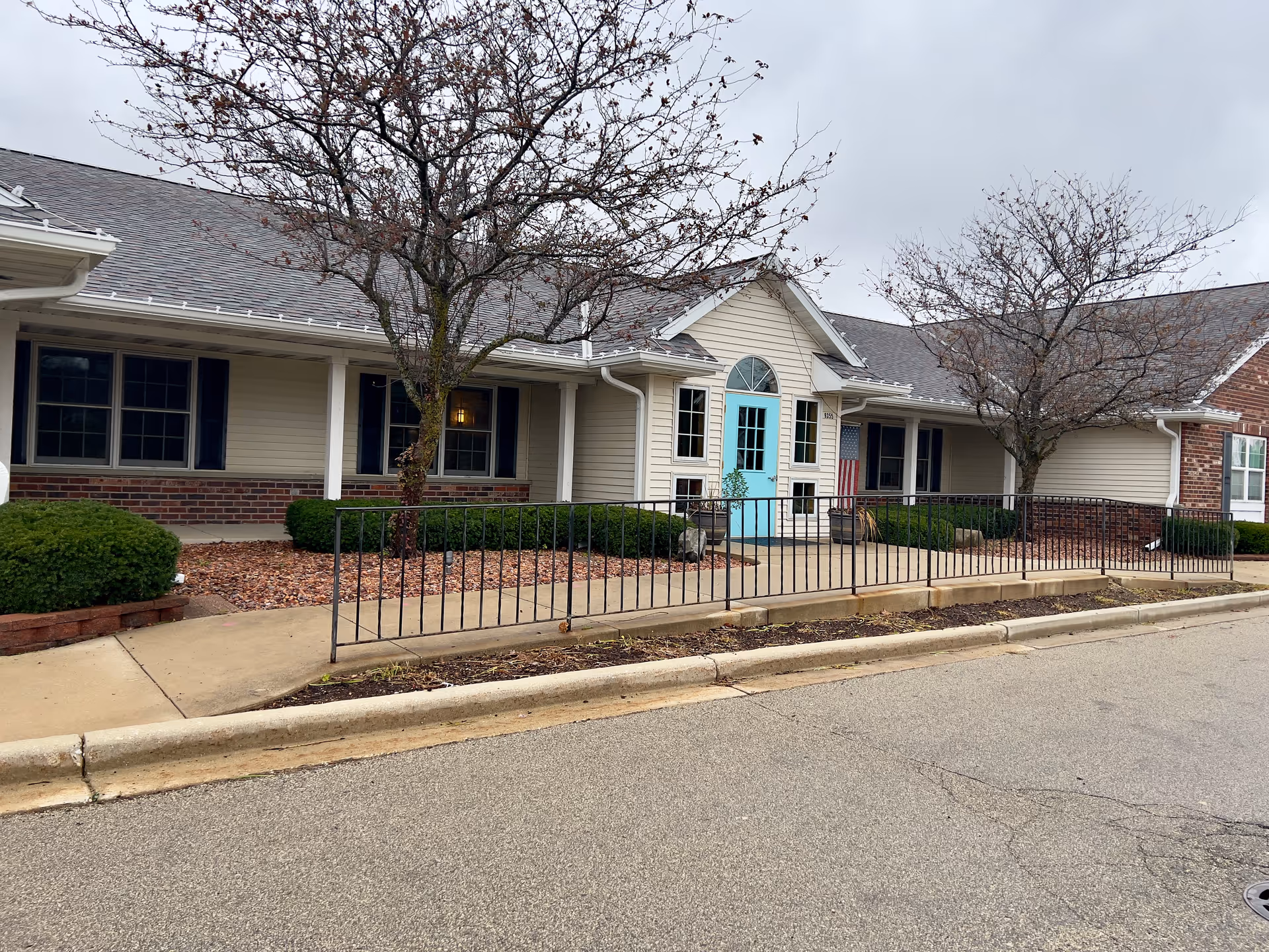 Exterior view of a single-story building with beige siding and brick accents. The building has a light blue door with windows on either side and a small porch area with a black metal railing. There are two leafless trees and some green shrubs in front of the building, under an overcast sky.