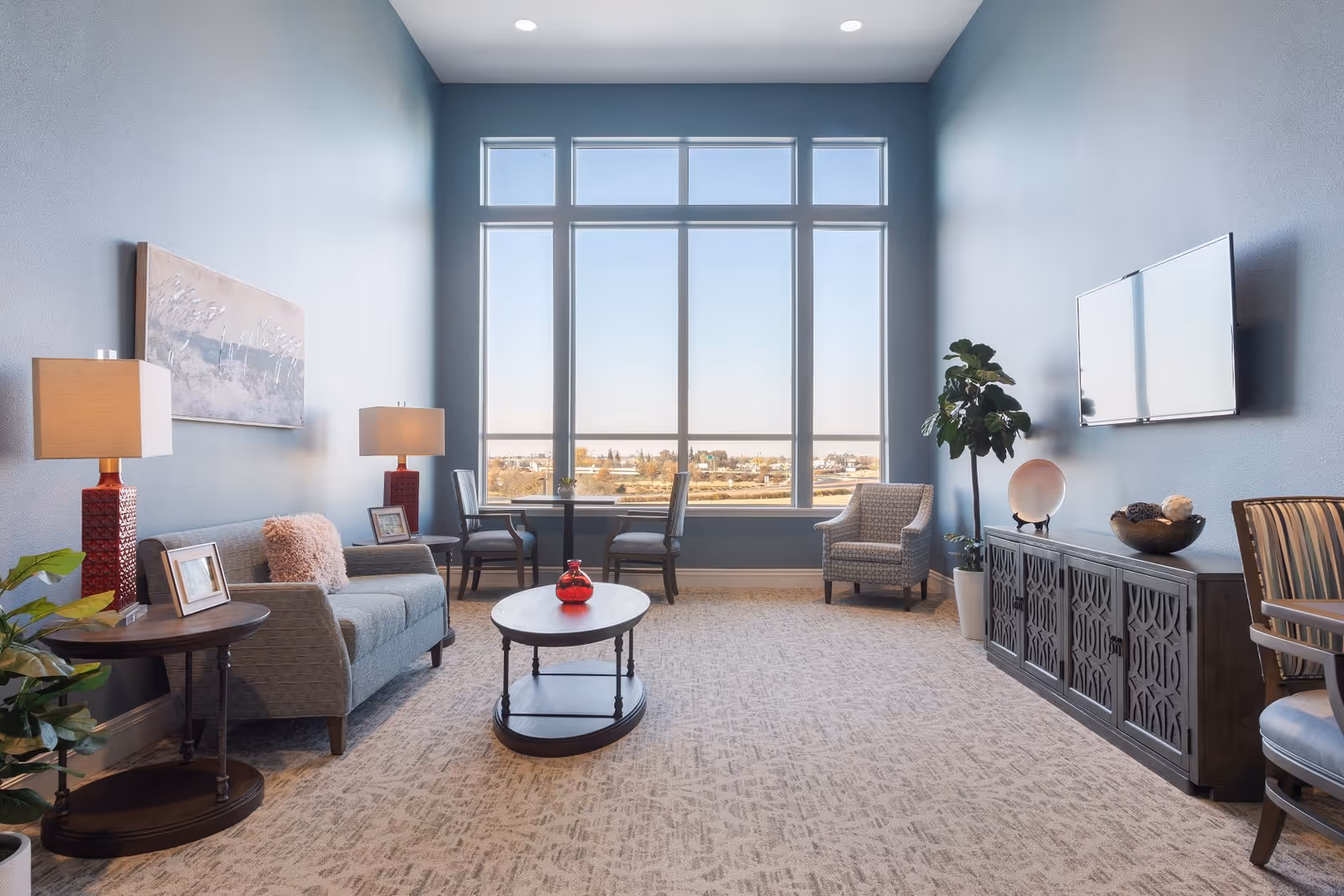 Bright lounge-style living room with a sofa, armchairs, a coffee table, a large floor-to-ceiling window and a wall-mounted TV.