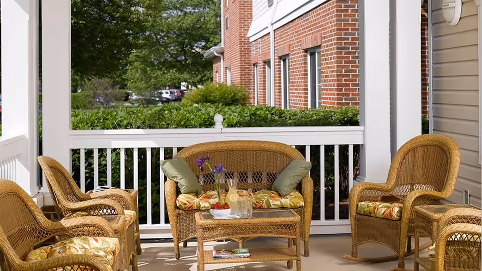 Outdoor patio area with wicker furniture including a loveseat, several chairs, and a coffee table. The furniture has floral and green cushions. There are plants and bushes along the white railing, and a brick building is visible in the background.