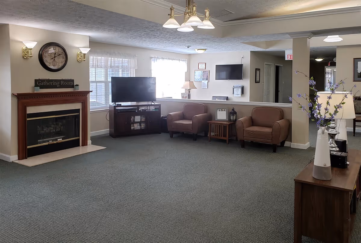 A cozy living room area with a fireplace on the left wall, a large wall clock above it, and a sign that reads 'Gathering Room'. There are two brown armchairs with a small wooden side table between them, a flat-screen TV on a dark wooden stand near the windows, and a vase with purple flowers on a wooden console table on the right. The room has carpeted flooring and soft lighting from wall sconces and ceiling fixtures.