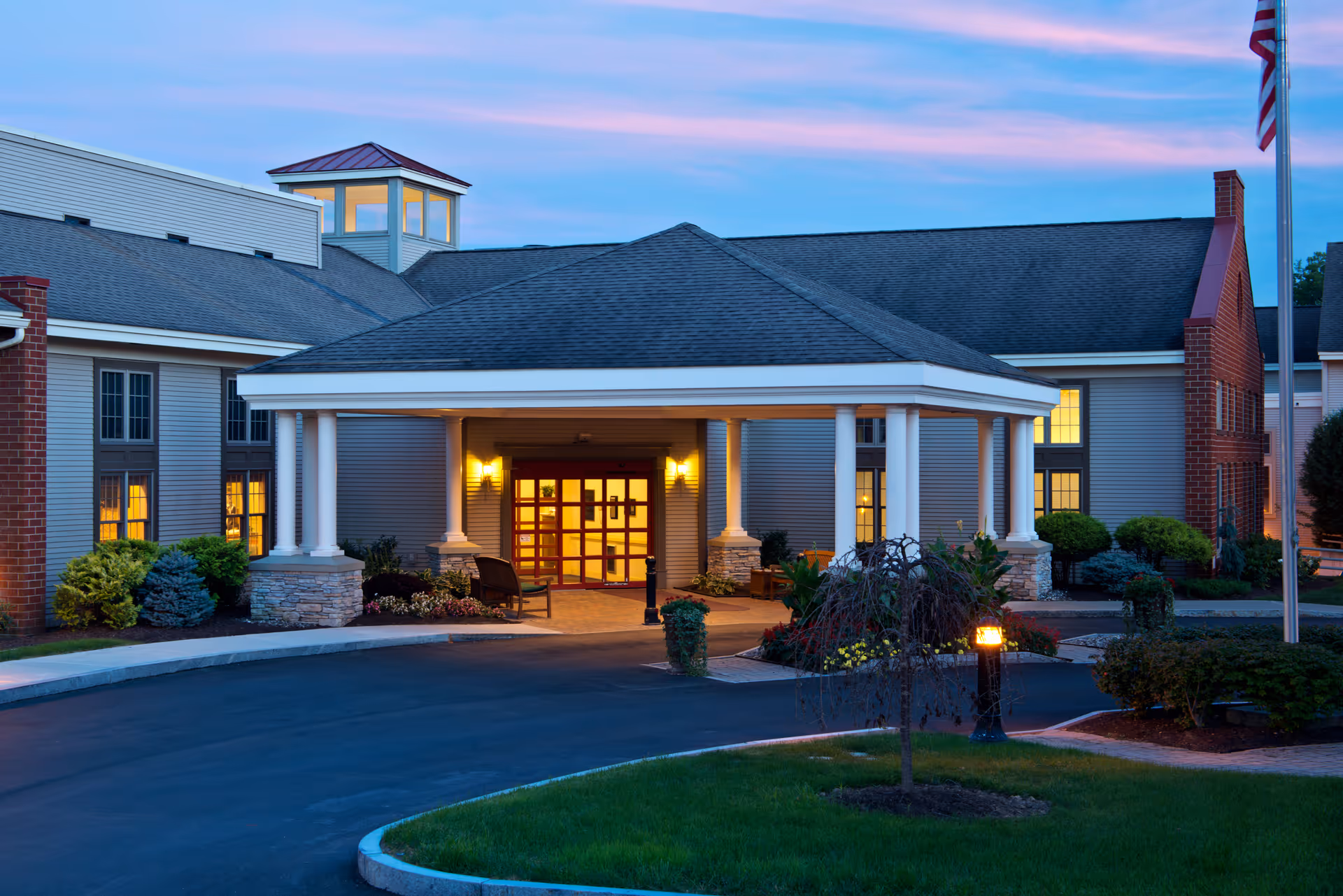 Exterior view of a senior living facility entrance at dusk with a covered drop-off area supported by white columns, well-maintained landscaping, and warm lights glowing from inside the building.