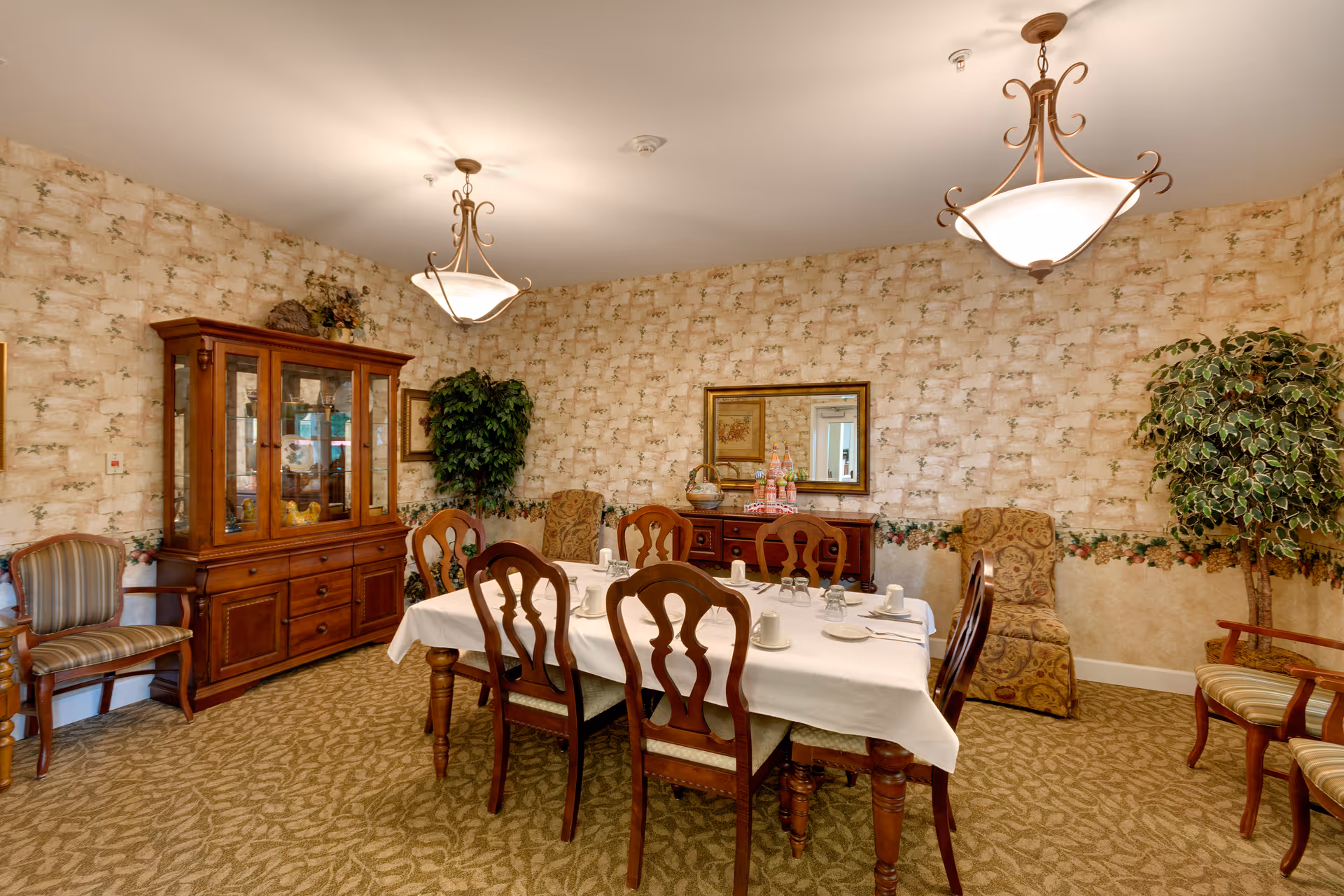 A dining room with a rectangular table covered with a white tablecloth set for six people with cups, saucers, and utensils. The room has patterned wallpaper, carpeted floor, two hanging light fixtures, a wooden china cabinet, a sideboard with a mirror above it, and several upholstered chairs and potted plants.