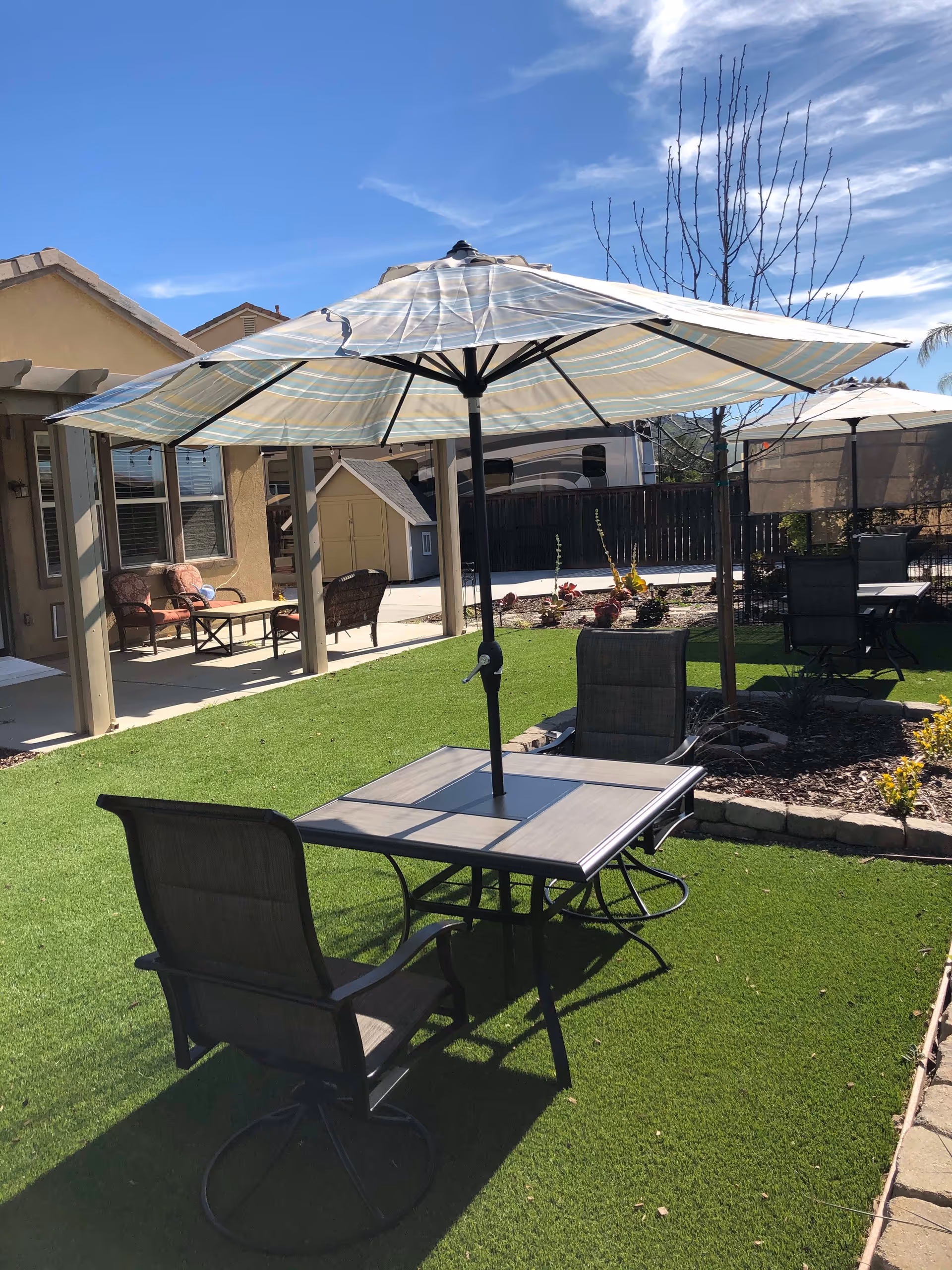 Outdoor seating area with a table and two chairs under a large striped umbrella on a green lawn. In the background, there is a patio with additional seating, a small shed, a wooden fence, and a clear blue sky with some clouds.