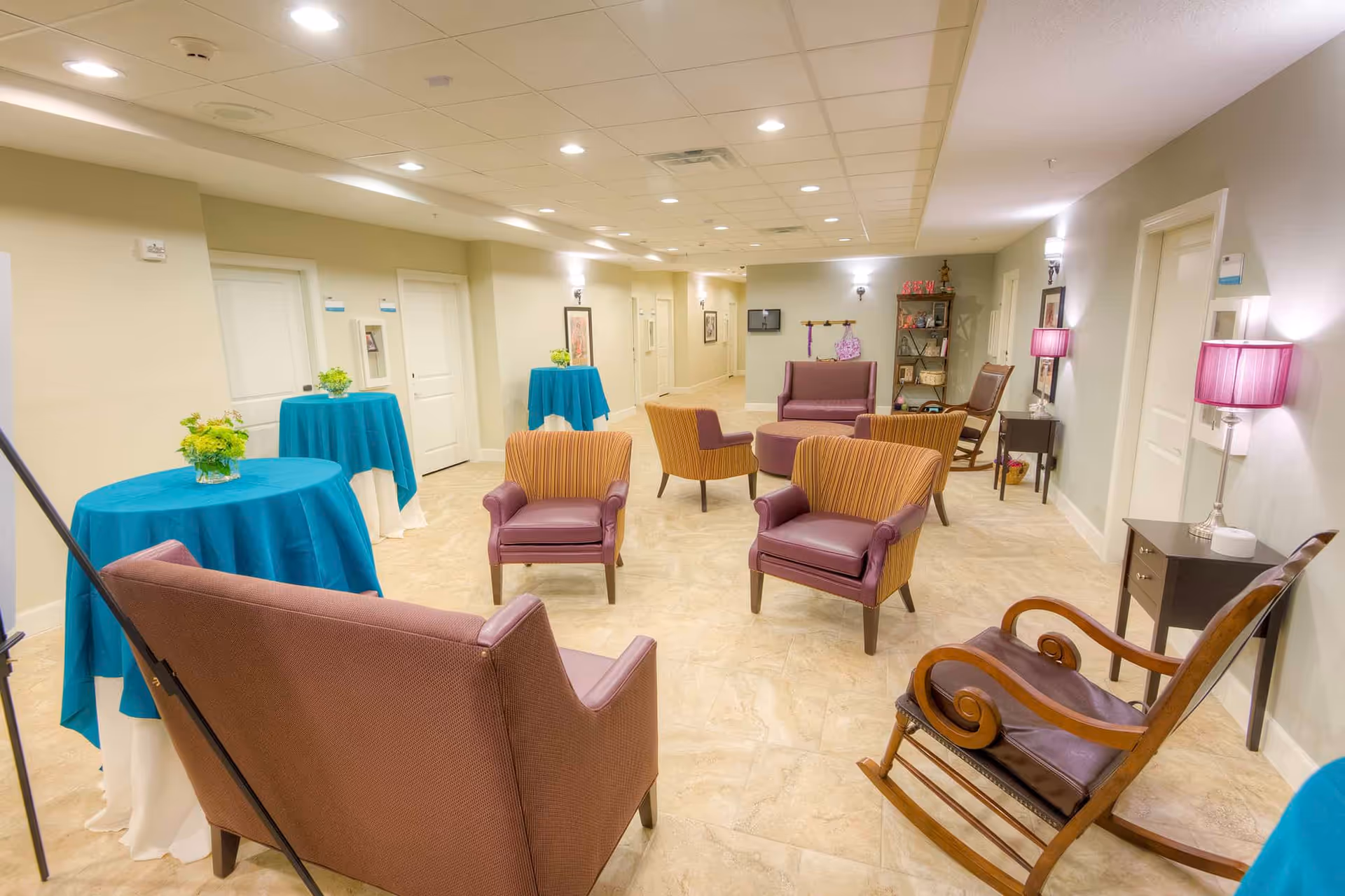 Communal lounge with upholstered chairs, a wooden rocking chair, and small round tables draped in teal tablecloths in a well-lit interior space.