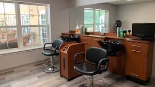 Interior view of a salon area in a senior living facility with two black salon chairs, a wooden cabinet with a sink for washing hair, a large mirror, and a window letting in natural light.