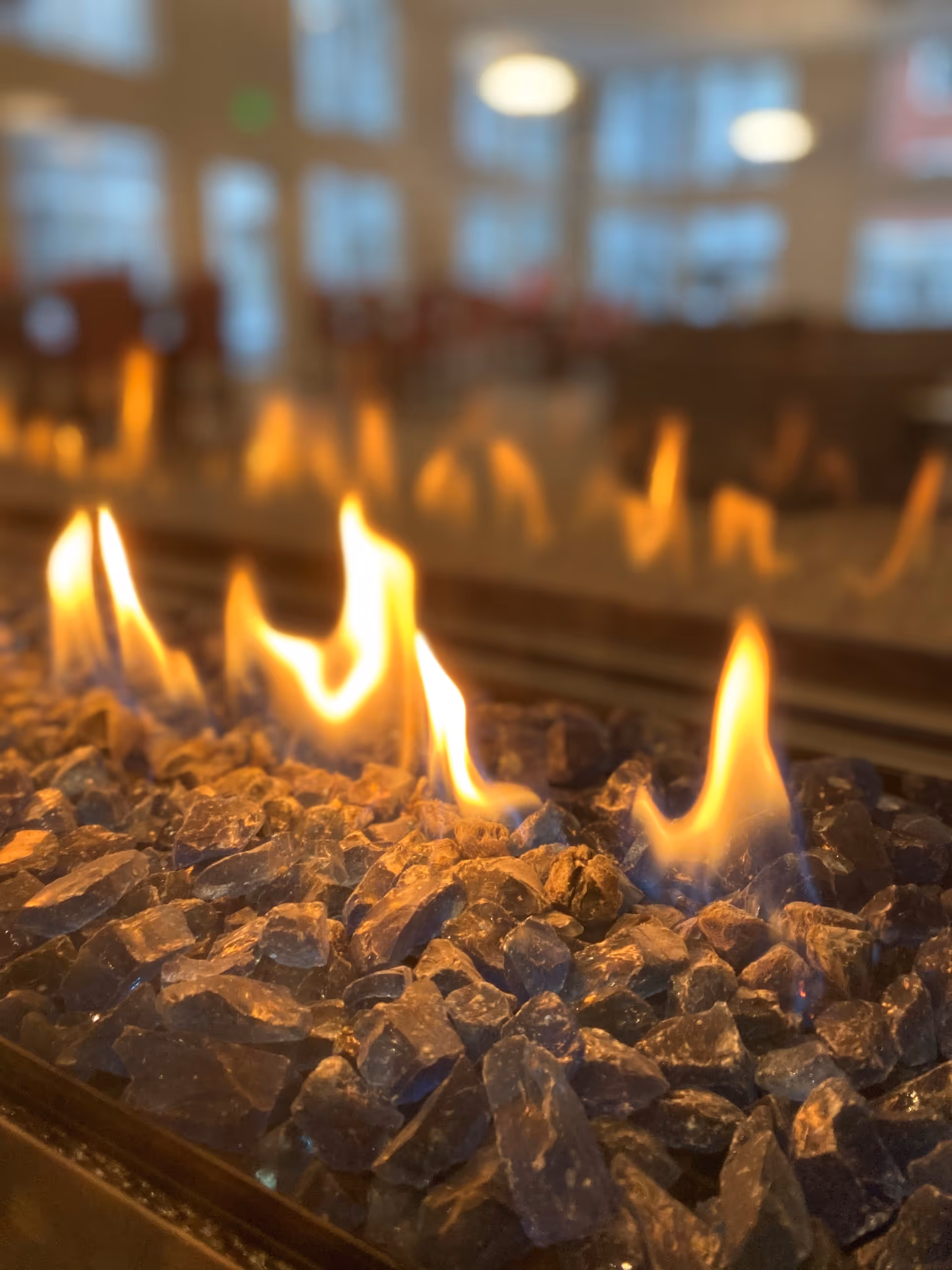 Close-up of flames dancing over blue fire glass in a modern indoor fireplace with a blurred seating area beyond.