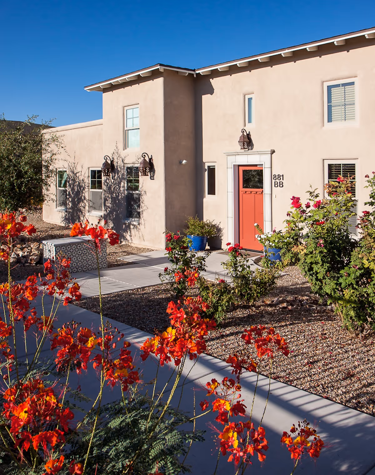 Front exterior of a beige stucco building with a red door and vibrant red flowers in the foreground.
