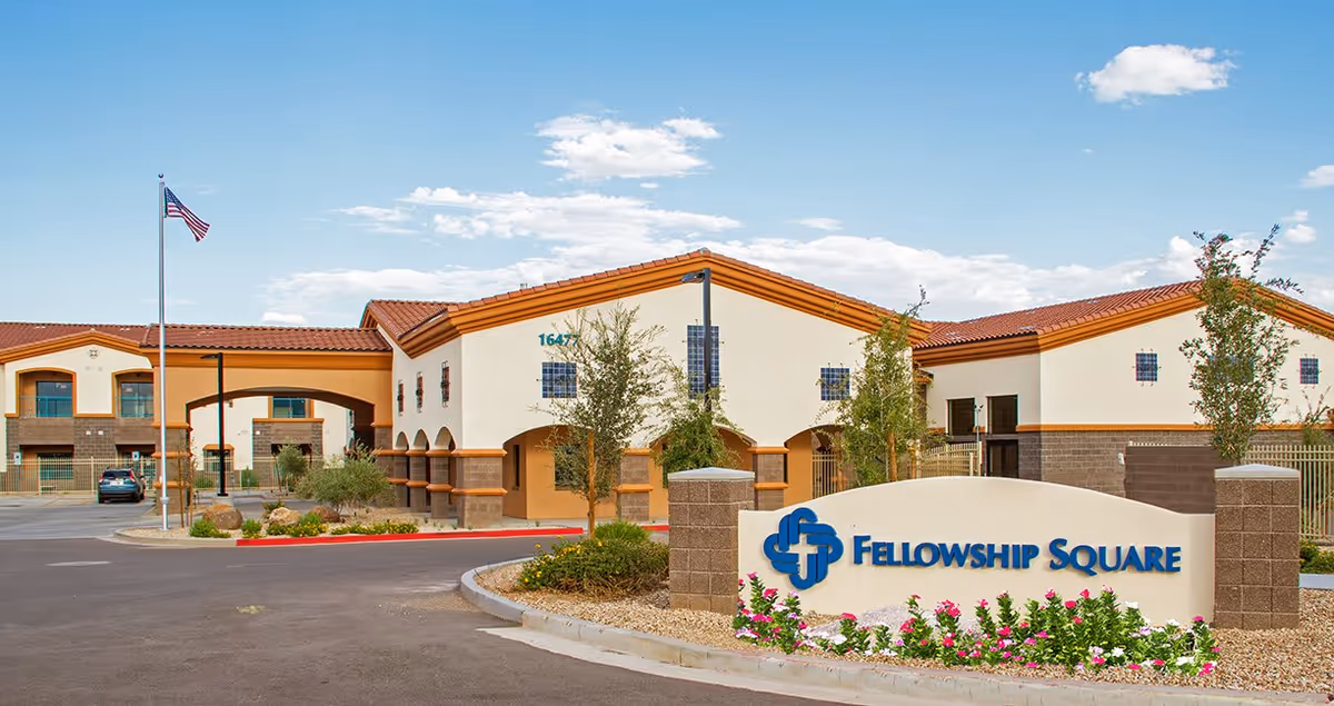 Exterior view of Fellowship Square Surprise senior living facility with a clear blue sky, an American flag on a flagpole, landscaped bushes and flowers, and a large sign displaying the facility's name in front.