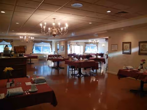 A spacious dining room in a senior facility with red tablecloths and chandeliers.