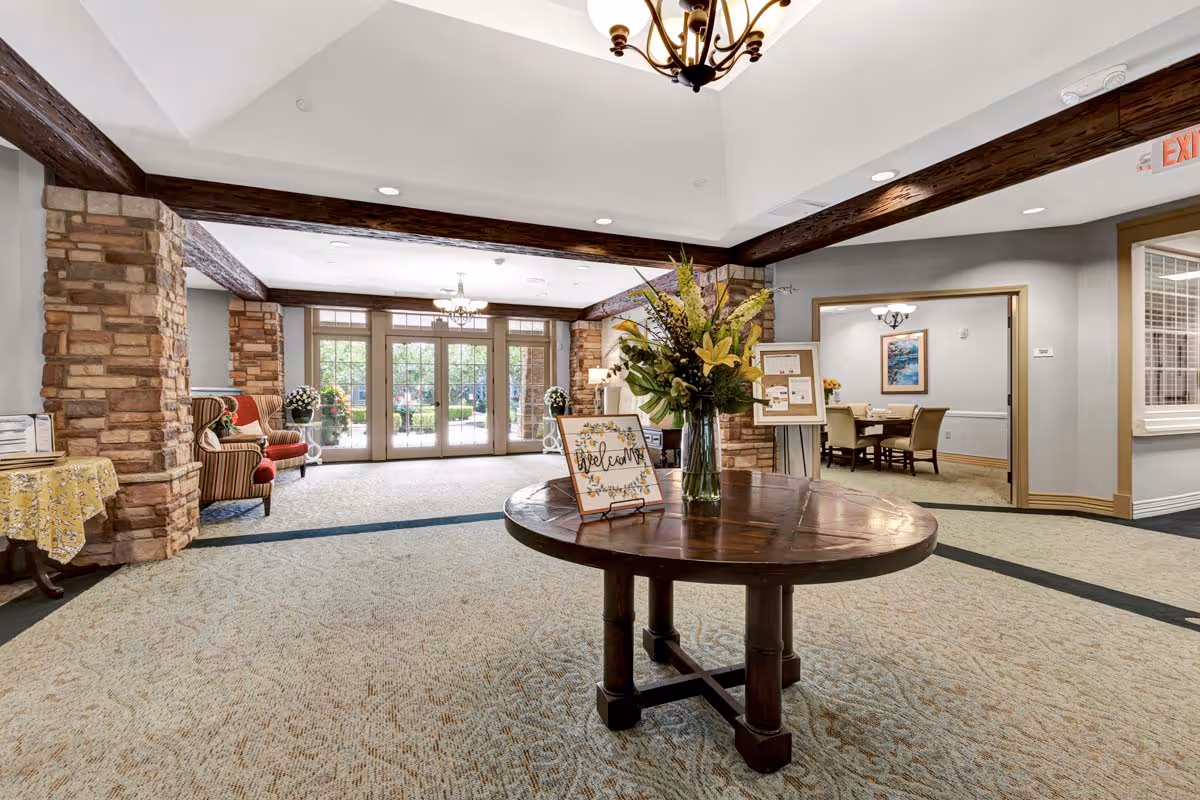 Spacious and well-lit lobby area with a round wooden table in the center holding a vase of flowers and a welcome sign. The room features stone pillars, wooden ceiling beams, carpeted floor, and large glass doors leading outside. Comfortable armchairs are placed along the walls, and an adjacent room with a dining table and chairs is visible.