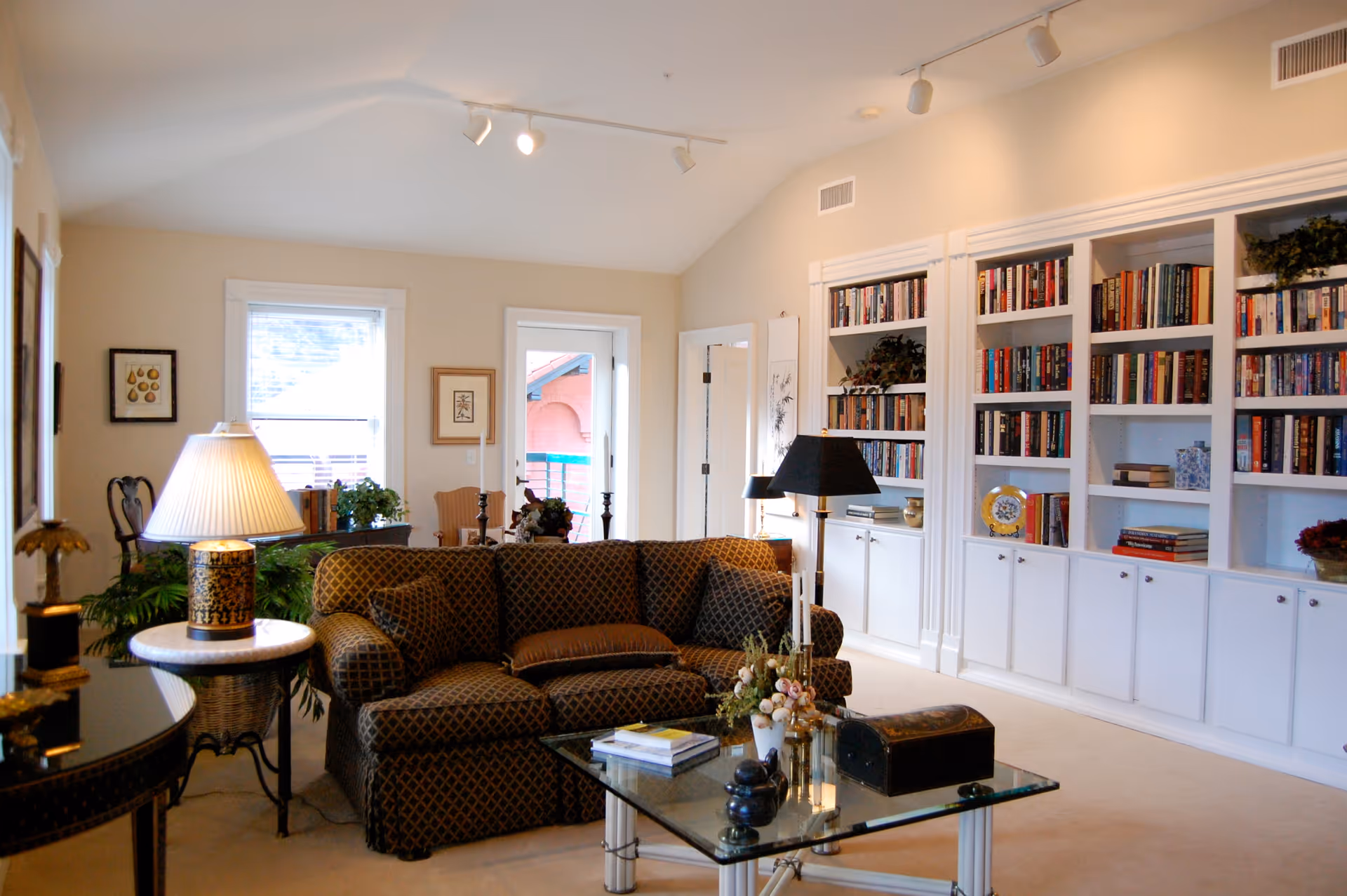 Cozy living room with a patterned sofa, glass coffee table, lamps, and built-in bookshelves filled with books.