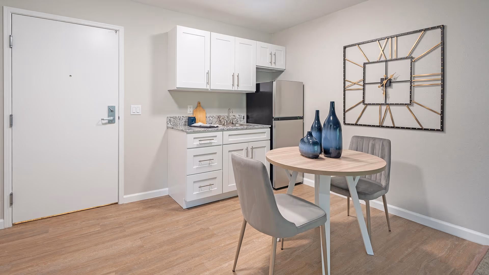 A small kitchen area with white cabinets, a granite countertop, a stainless steel refrigerator, and a sink. In front of the kitchen is a round wooden table with two gray upholstered chairs. On the table are three decorative blue vases. A large wall clock with Roman numerals is mounted on the wall behind the table. The floor is wood, and a white door is visible to the left.