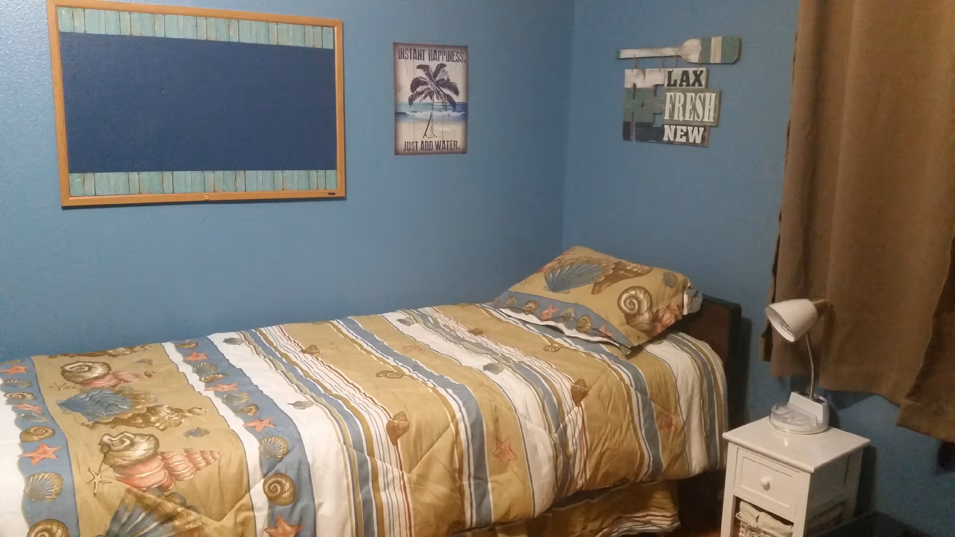 A small bedroom with blue walls featuring a single bed with a seashell-themed bedspread and pillow. Above the bed are three wall decorations, including a framed corkboard, a sign with a palm tree and the text 'INSTANT HAPPINESS JUST ADD WATER,' and a wooden sign with the words 'RELAX FRESH NEW.' Next to the bed is a white nightstand with a small white lamp and some items on top. A brown curtain covers the window on the right side.