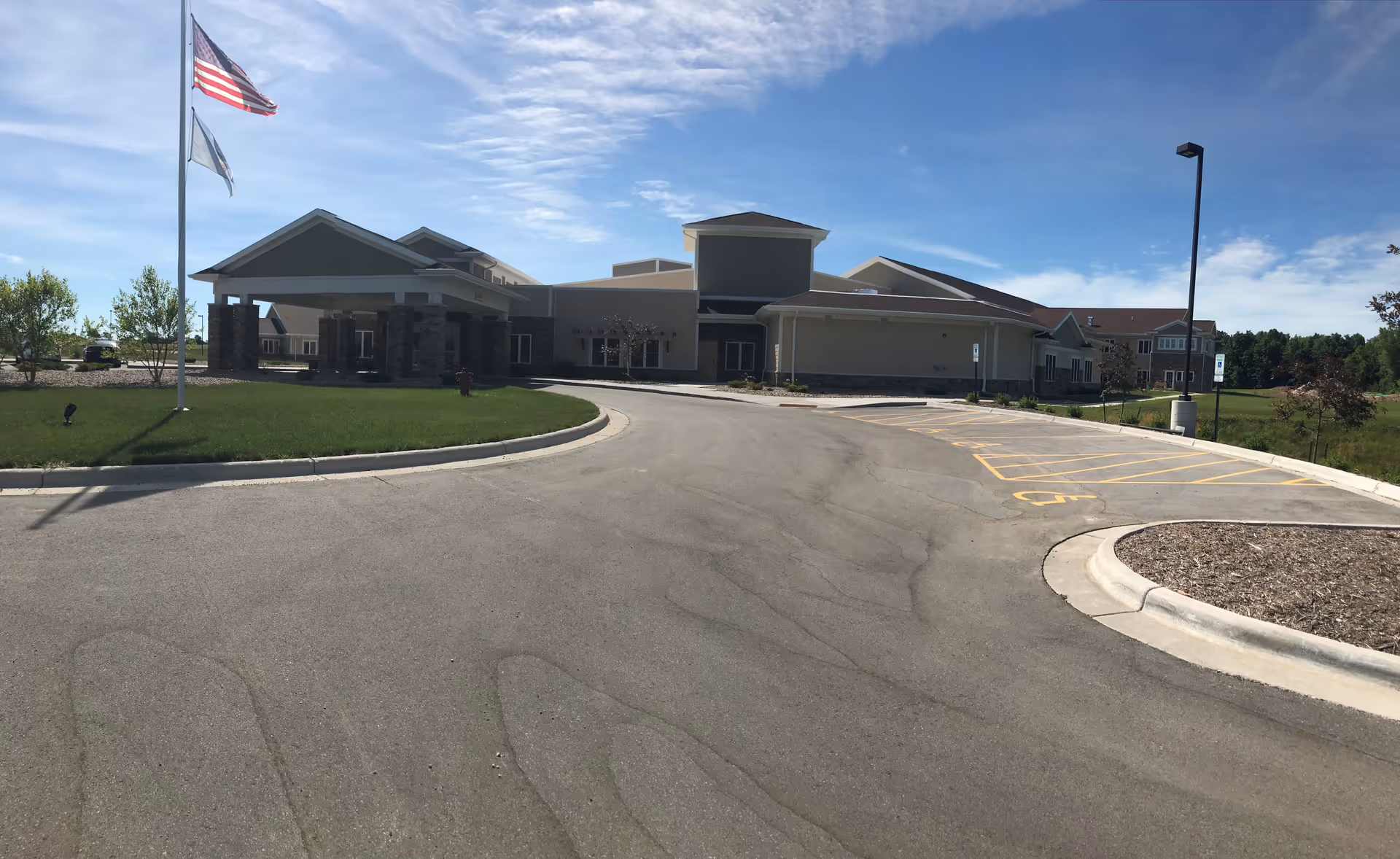 Exterior view of Oak Park Place Green Bay facility showing the main entrance with a covered drop-off area, a flagpole with the American flag and another flag, a parking area with handicap spaces, and a clear blue sky.