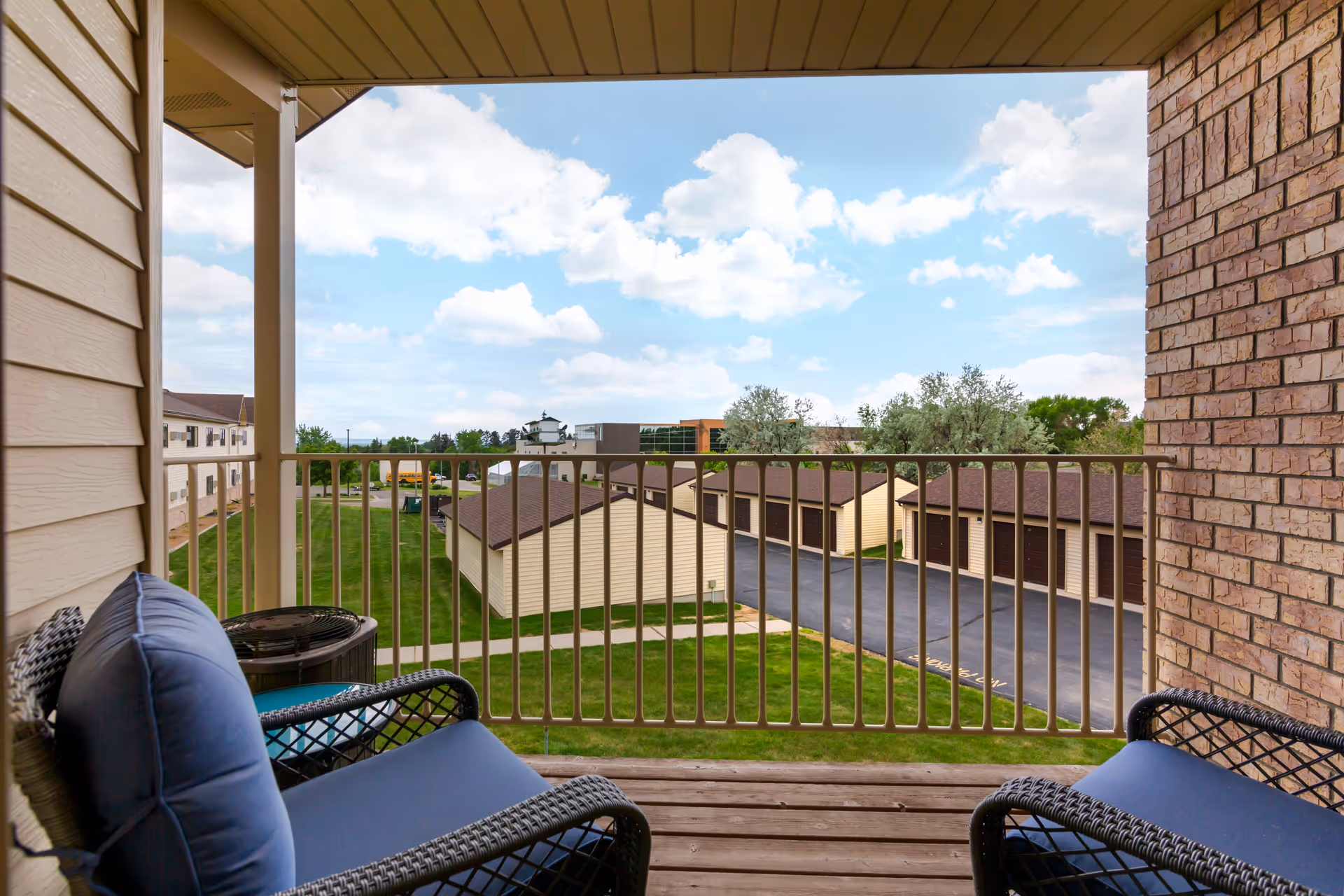 View from a covered balcony with two cushioned wicker chairs facing outward. The balcony overlooks a grassy area, a row of garages with brown doors, and a partly cloudy blue sky.
