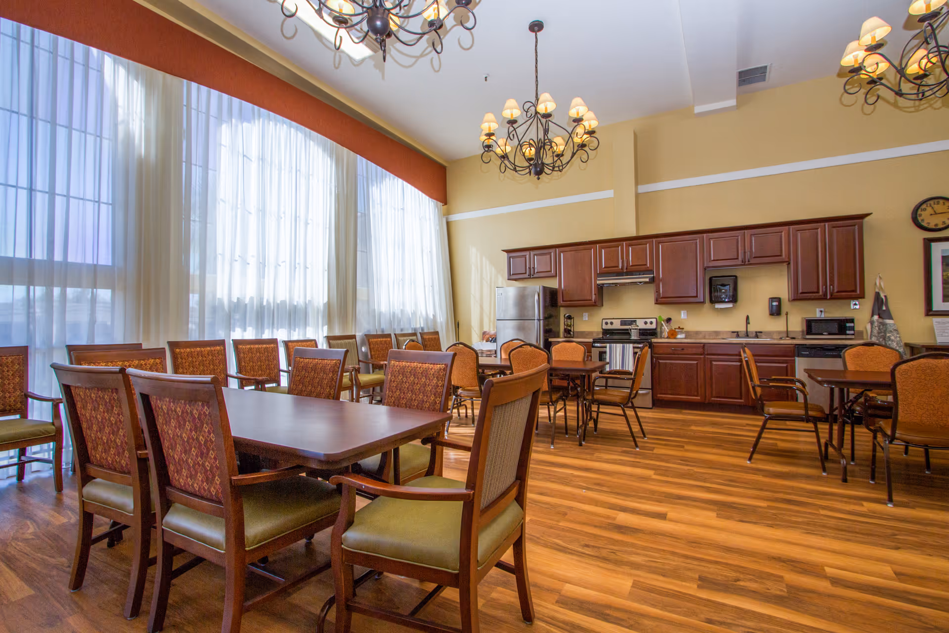 A spacious dining room with wooden tables and chairs arranged neatly. The room features large windows with sheer white curtains allowing natural light to fill the space. There are two ornate chandeliers hanging from the ceiling. In the background, there is a kitchen area with wooden cabinets, a stainless steel refrigerator, stove, microwave, and a sink. The floor is made of wood, and the walls are painted a warm beige color.