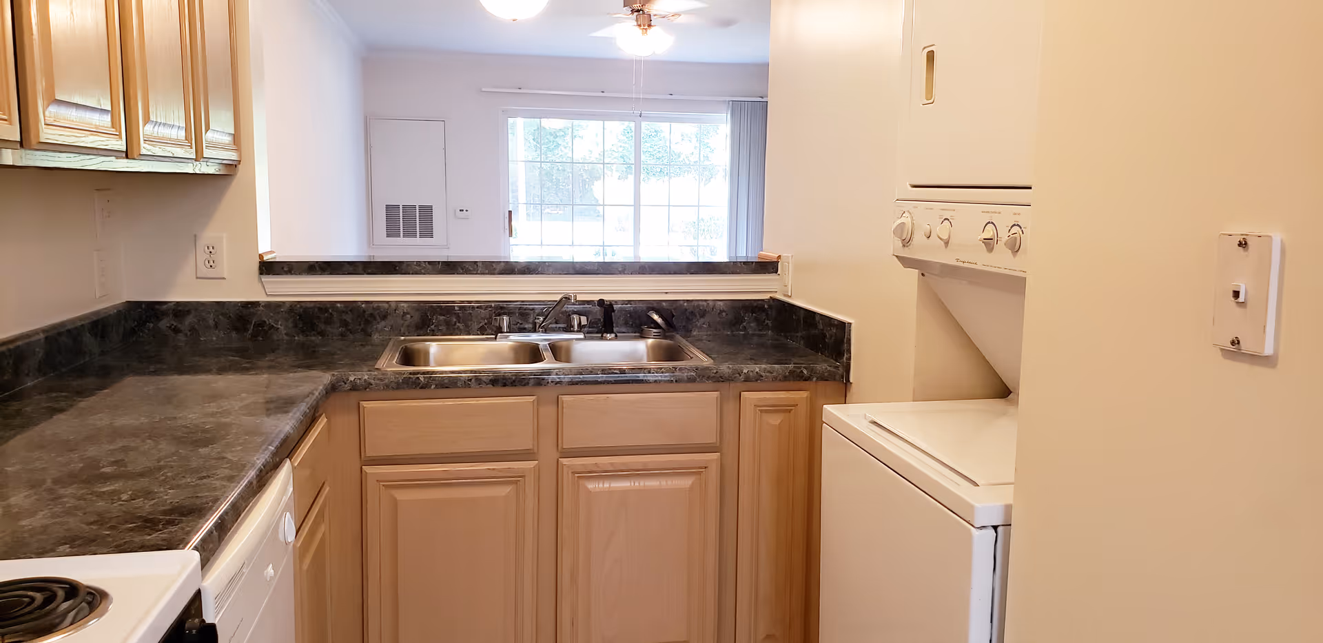 Small apartment kitchen with dark marble-look countertops, a double sink, light wood cabinets, and a stacked washer/dryer opening to the living room.