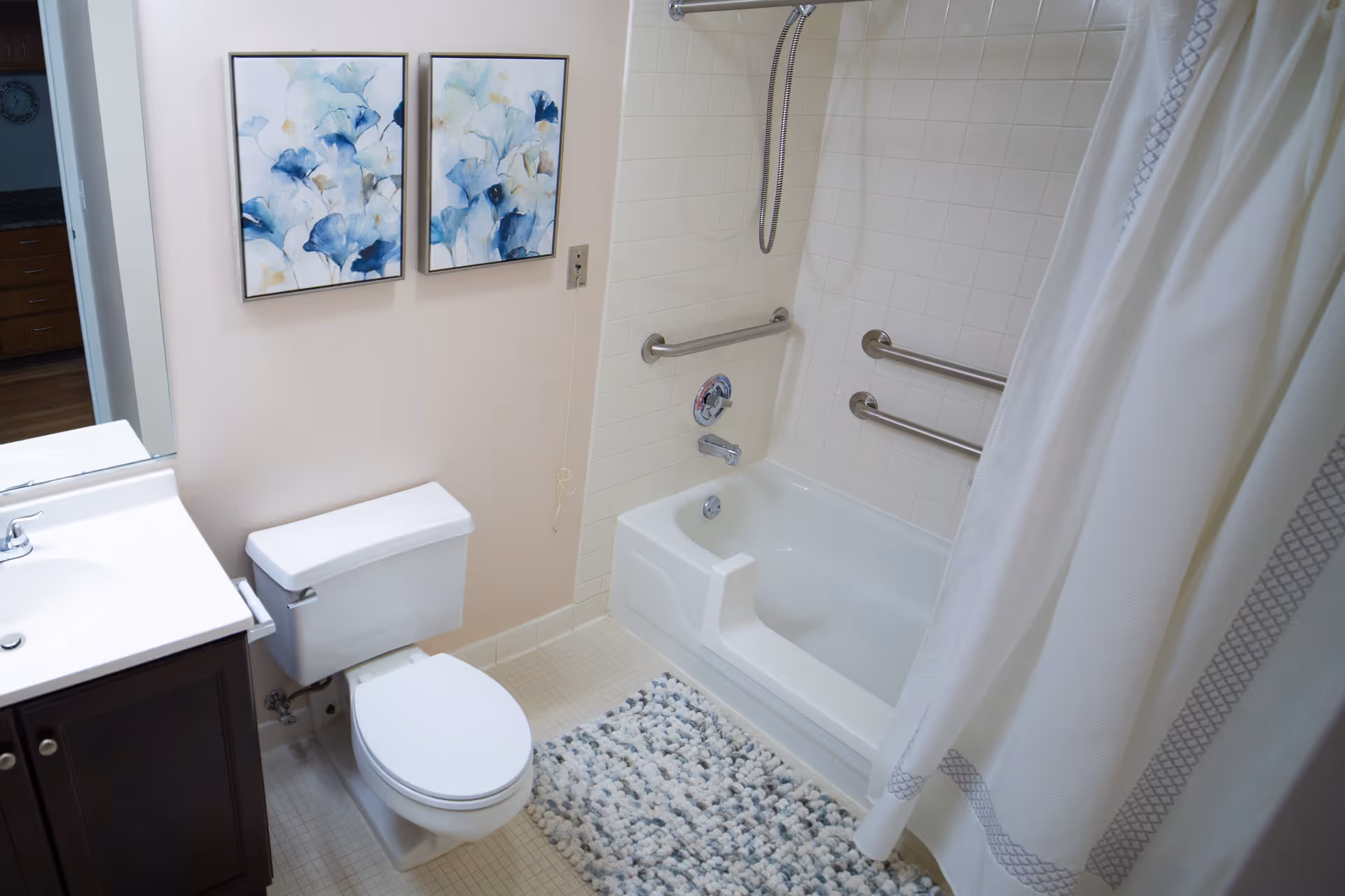 A clean bathroom featuring a white toilet, a white sink with a dark wooden cabinet, a bathtub with grab bars and a handheld showerhead, a white shower curtain with a gray pattern, a textured bath mat, and two framed floral paintings on the wall.