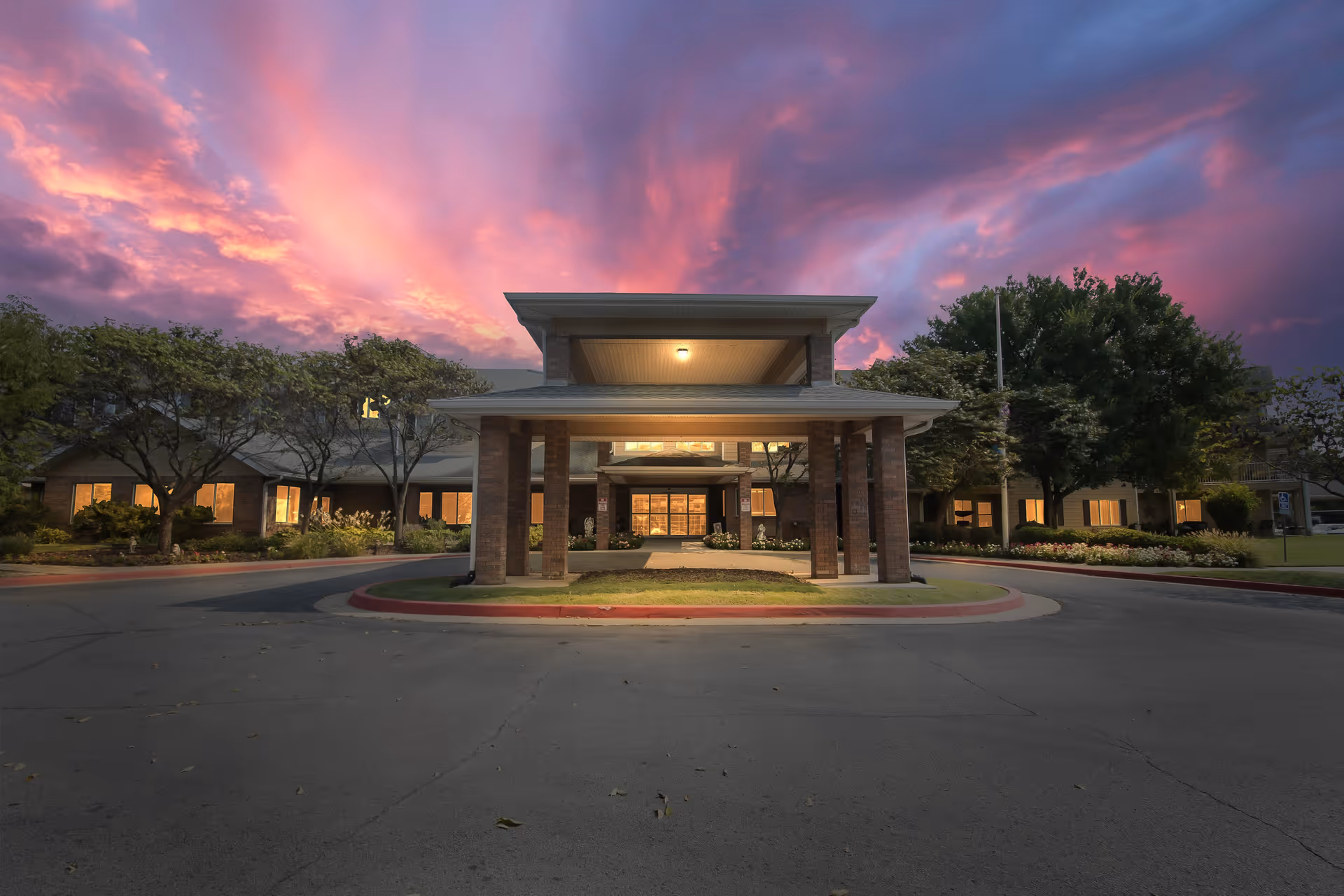 Front exterior view of Morada Broken Arrow facility at dusk with a covered entrance, illuminated windows, surrounding trees, and a dramatic pink and purple sky.