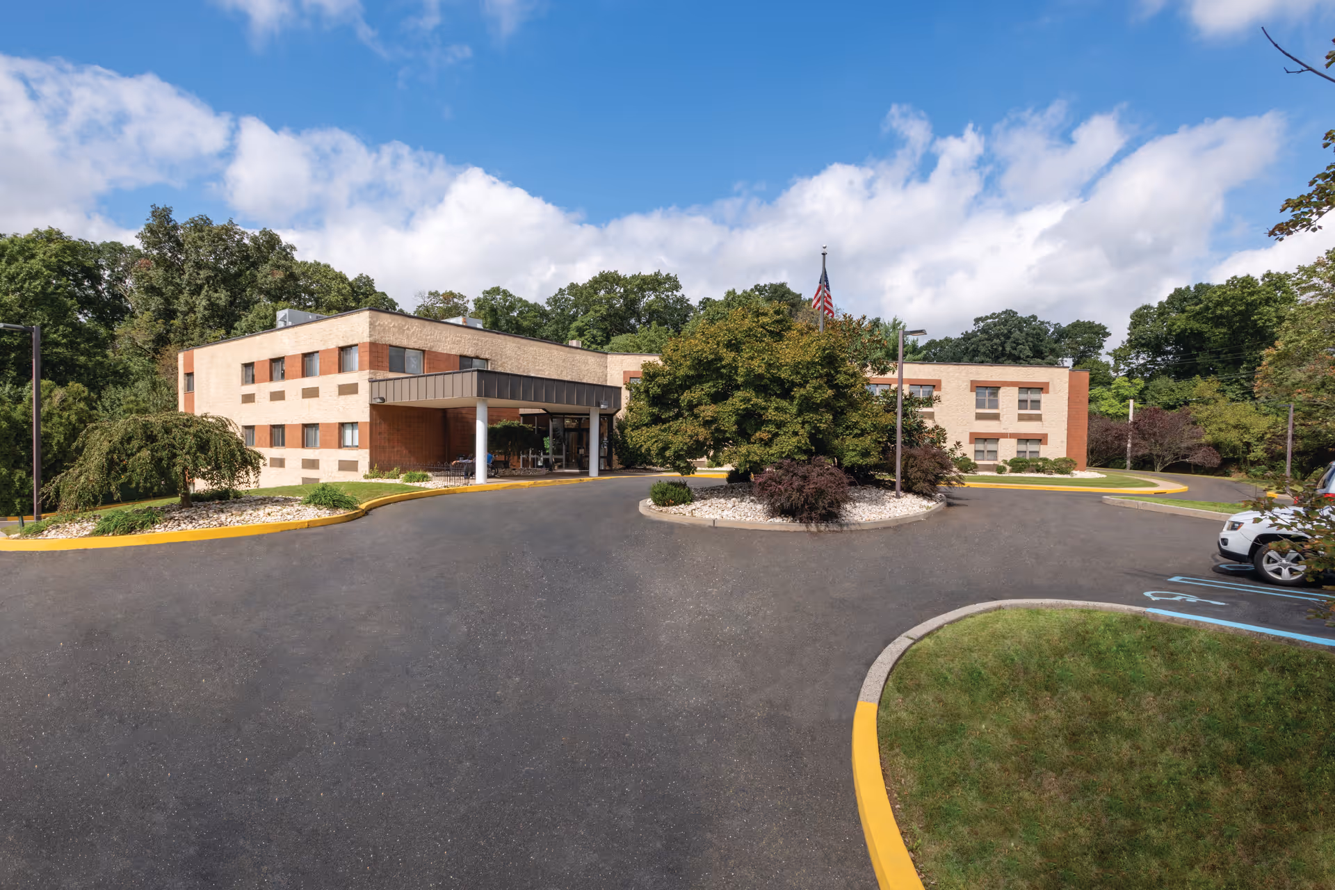 Exterior view of Dresher Hill Health & Rehabilitation Center, a two-story brick building with a covered entrance, surrounded by trees and landscaping under a partly cloudy sky.