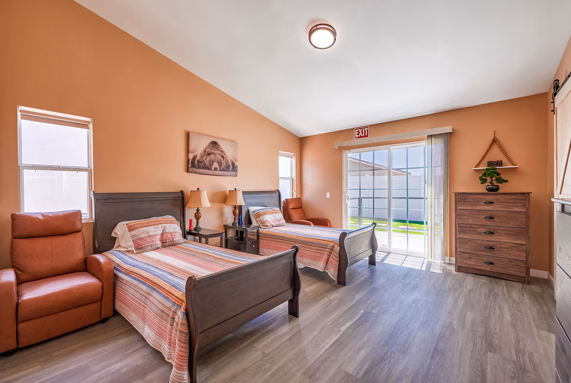A bright bedroom with two single beds featuring wooden headboards and striped bedspreads. Between the beds are two small nightstands with lamps. There are two windows on the left wall and a sliding glass door on the far wall with an exit sign above it. To the right is a wooden dresser with a small plant on top and a decorative shelf above it. The walls are painted a warm peach color and the floor is light wood.