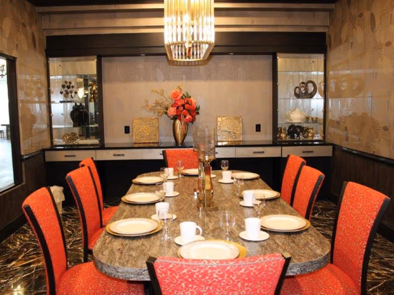 A dining room with a large rectangular marble table set with white plates, cups, and glassware. The table is surrounded by red upholstered chairs. A decorative vase with flowers is placed at the center of the table. The room features patterned wallpaper, a chandelier above the table, and built-in cabinets with glass display shelves on the back wall.